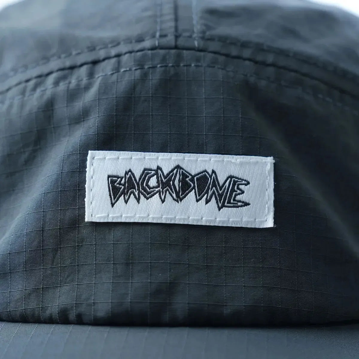 Close-up of the Backbone Ride Club Hat in dark gray, featuring a rectangular white patch with “BACKBONE” in stylized black lettering.