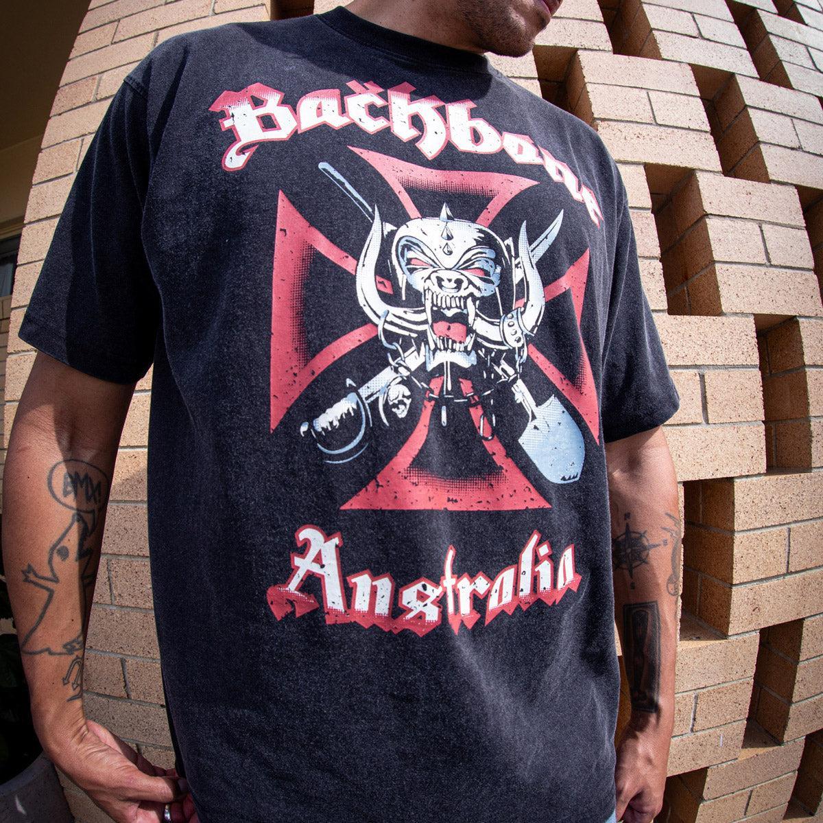 A person wears a black Backbone Road Crew Vintage T-Shirt with a horned skull graphic, standing in front of a brick wall and showcasing this premium cotton heavyweight tee.