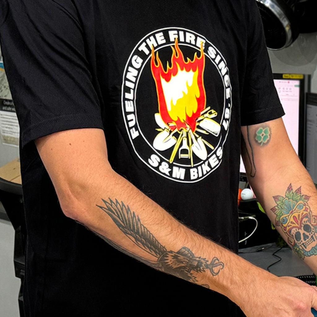 A tattooed person wears the S&M Fuel The Fire T-Shirt, a black tee with a flaming logo and "FUELING THE FIRE SINCE '87 S&M BIKES," printed in California.