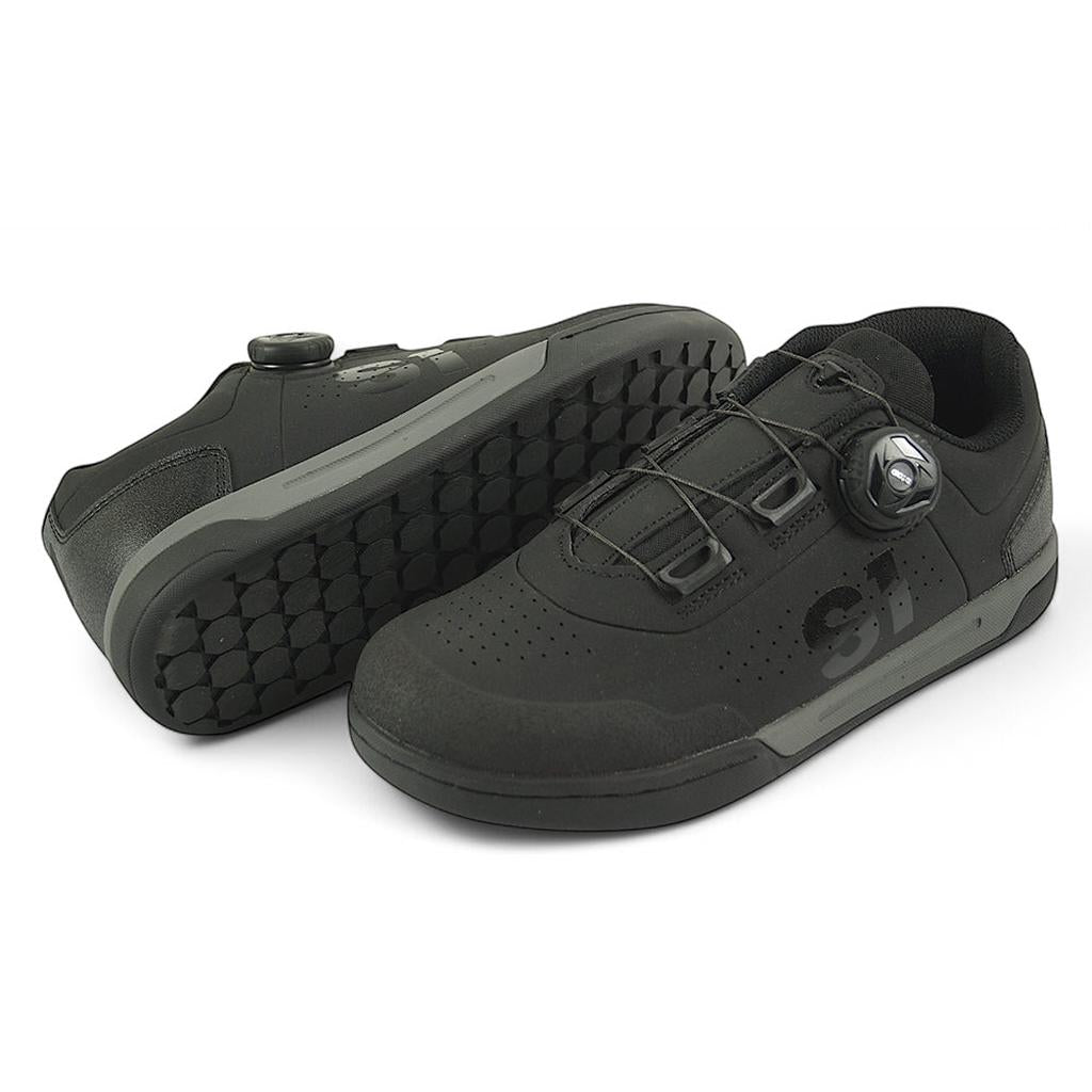 A pair of S1 Accel Race Shoes in black featuring textured, impact-resistant soles for ultimate grip, perforated toes, and a dial-based lacing system. One shoe stands upright while the other lies sideways displaying the sole.