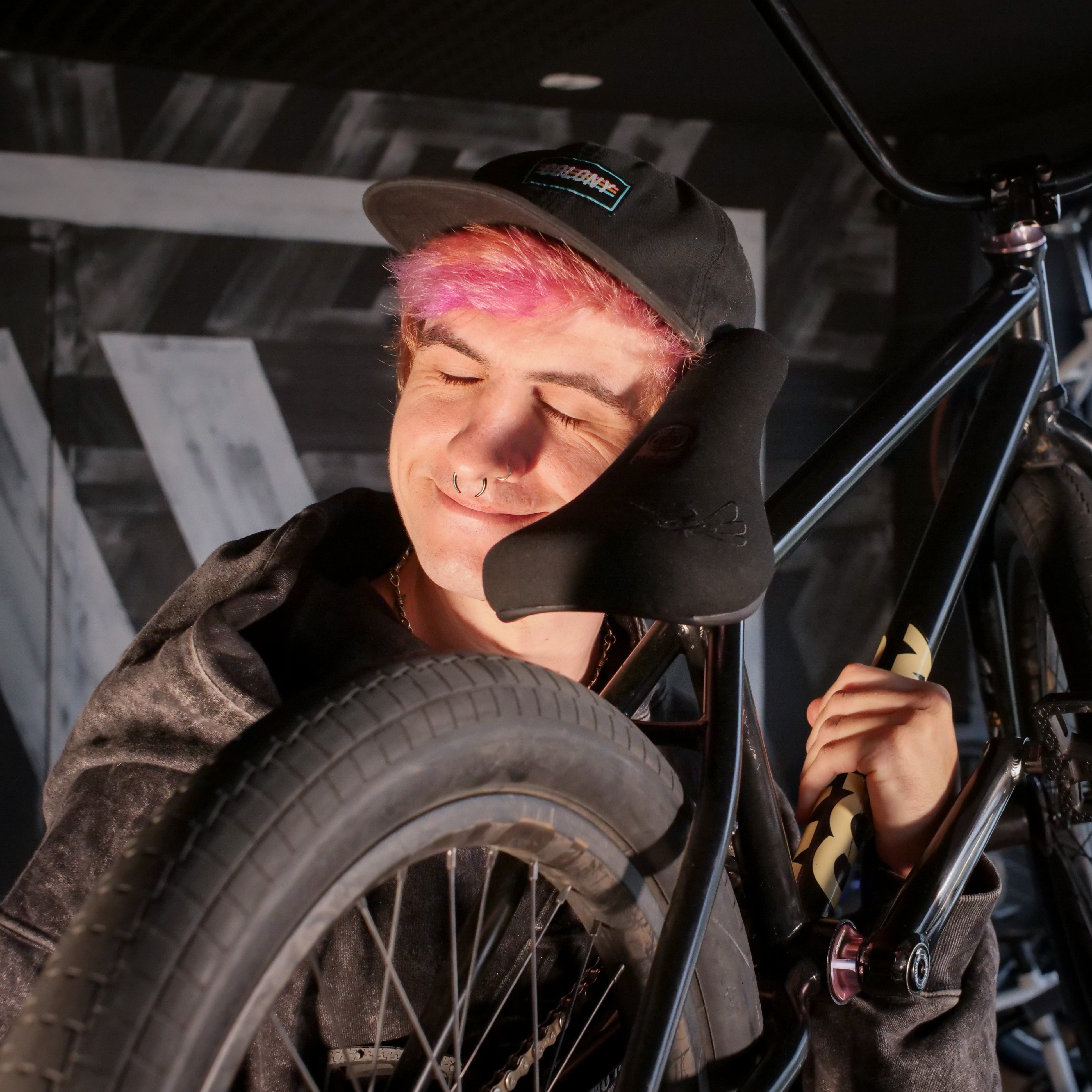 A person with pink hair and a cap smiles with eyes closed, hugging the Colony Sigil Pivotal Seat (Sean Gardner Signature) BMX seat, indoors against dark patterned walls.