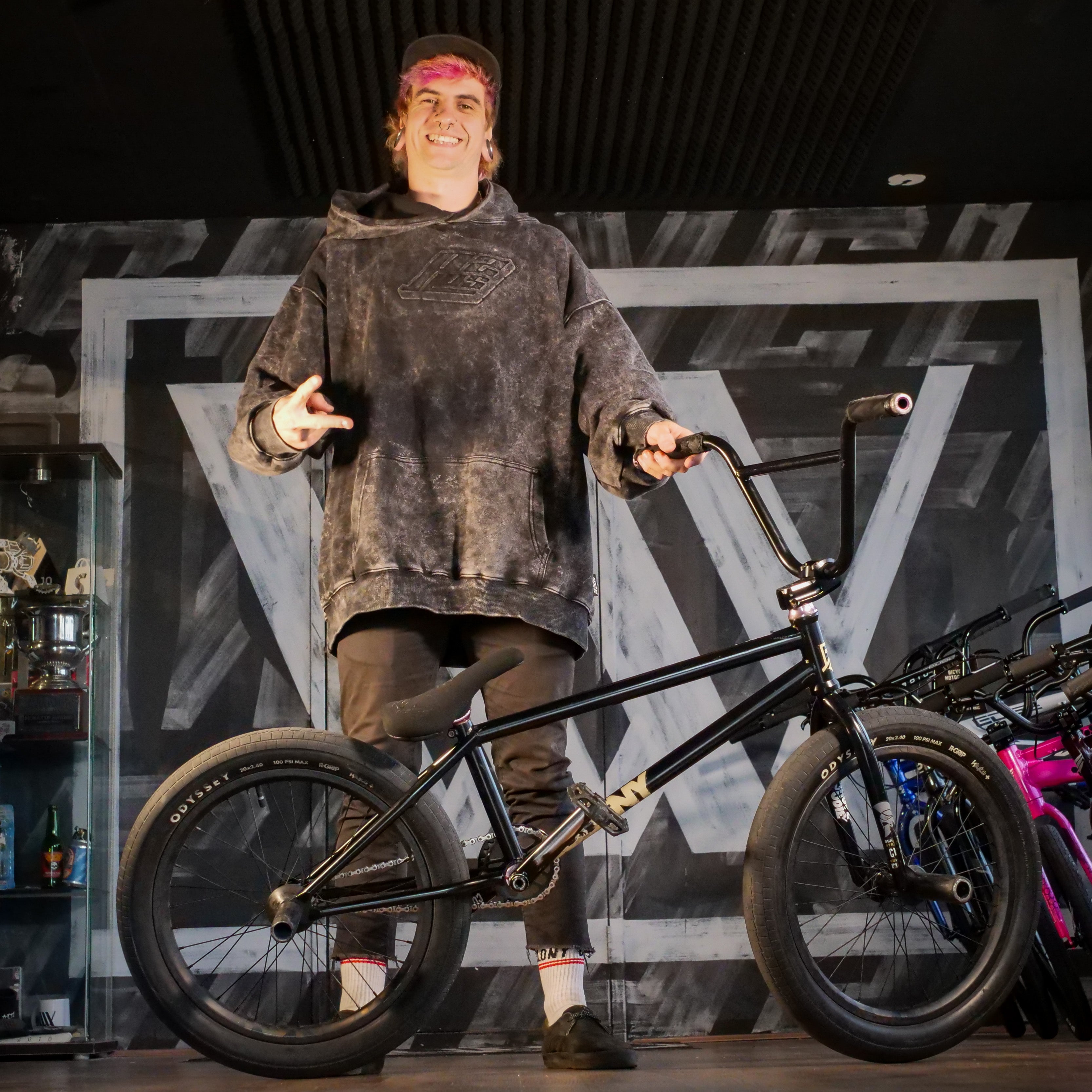 A person in a black hoodie stands indoors, smiling and making a peace sign while holding a black BMX bike featuring the Colony Sigil Pivotal Seat (Sean Gardner Signature).