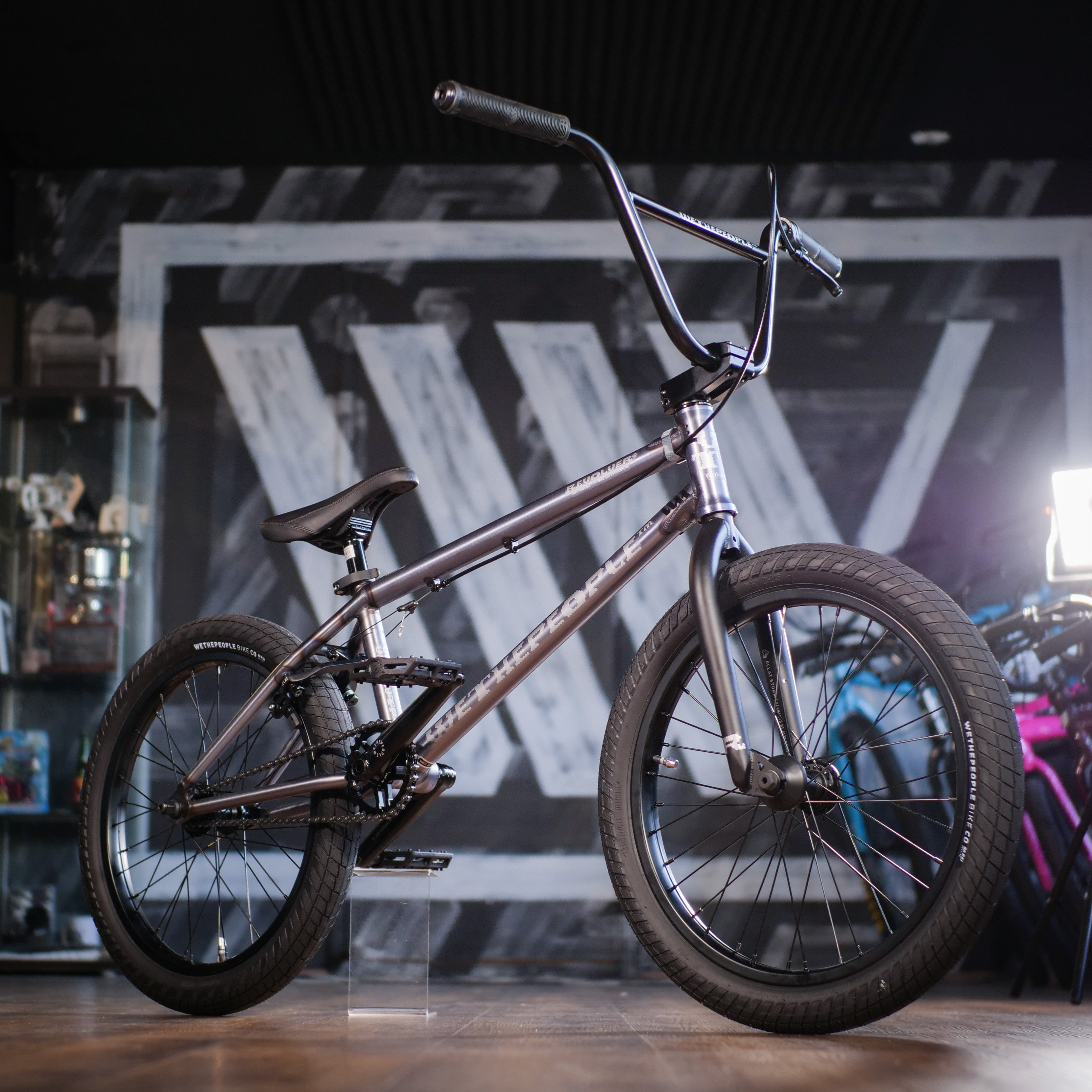 The Wethepeople Revolver 20 Inch Bike, featuring a 21" top tube and metallic frame with black handlebars, is displayed indoors on a wooden floor against a geometric mural backdrop.
