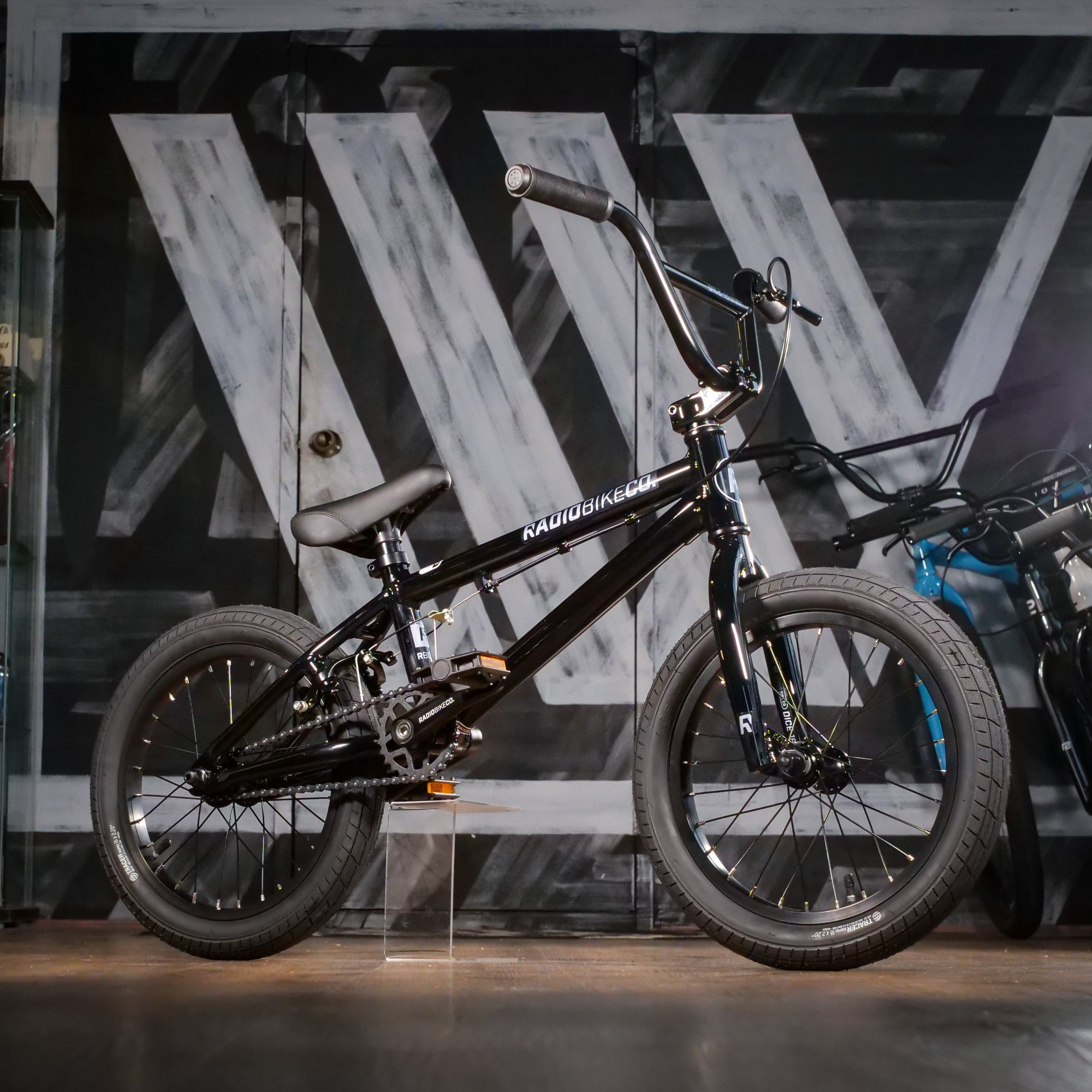 The Radio Dice 16 Inch Bike in black is shown indoors against a monochrome, industrial wall—a stylish and ideal 16 inch bike for kids.
