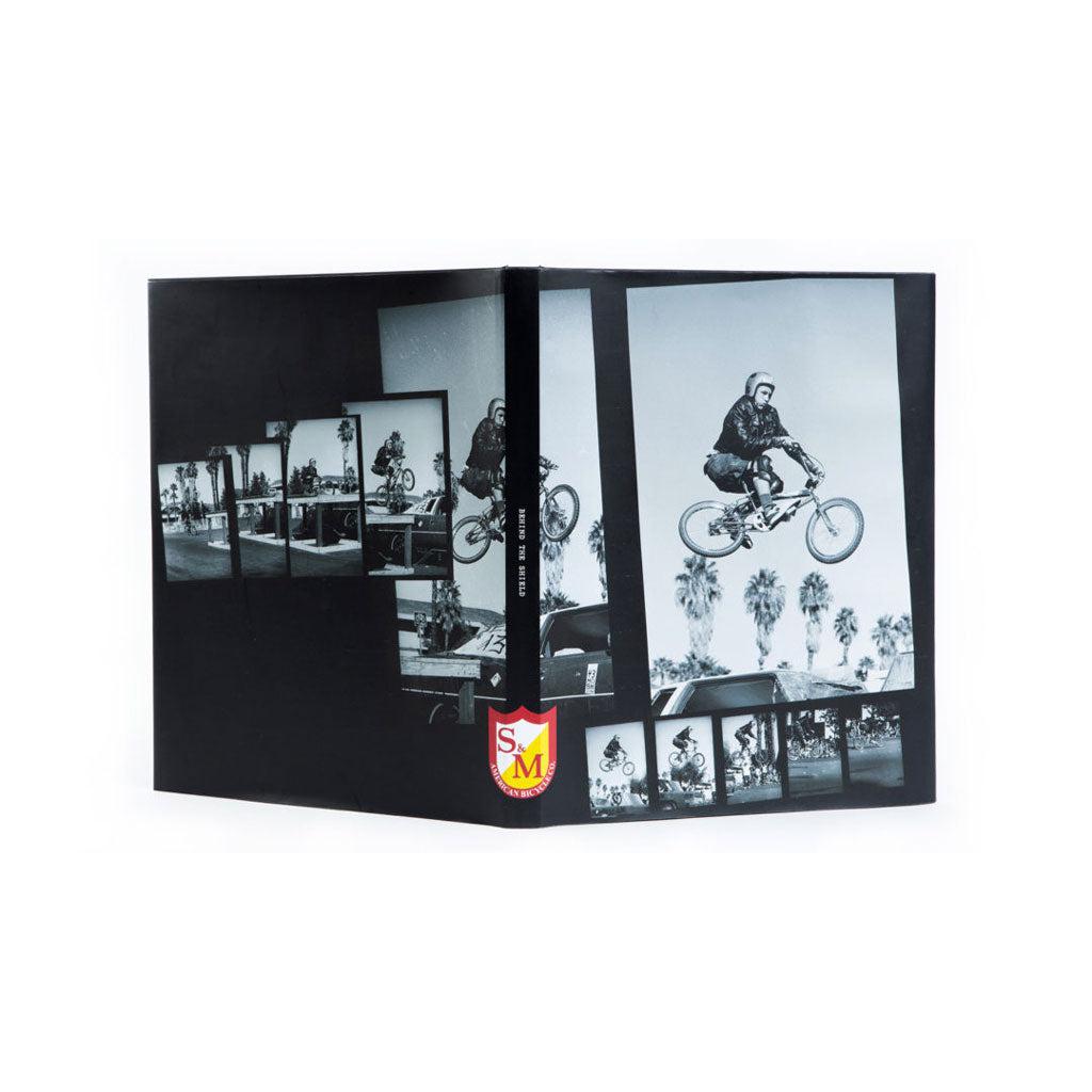 A black and white photo of a man riding the S&M Behind The Shield Book bike.