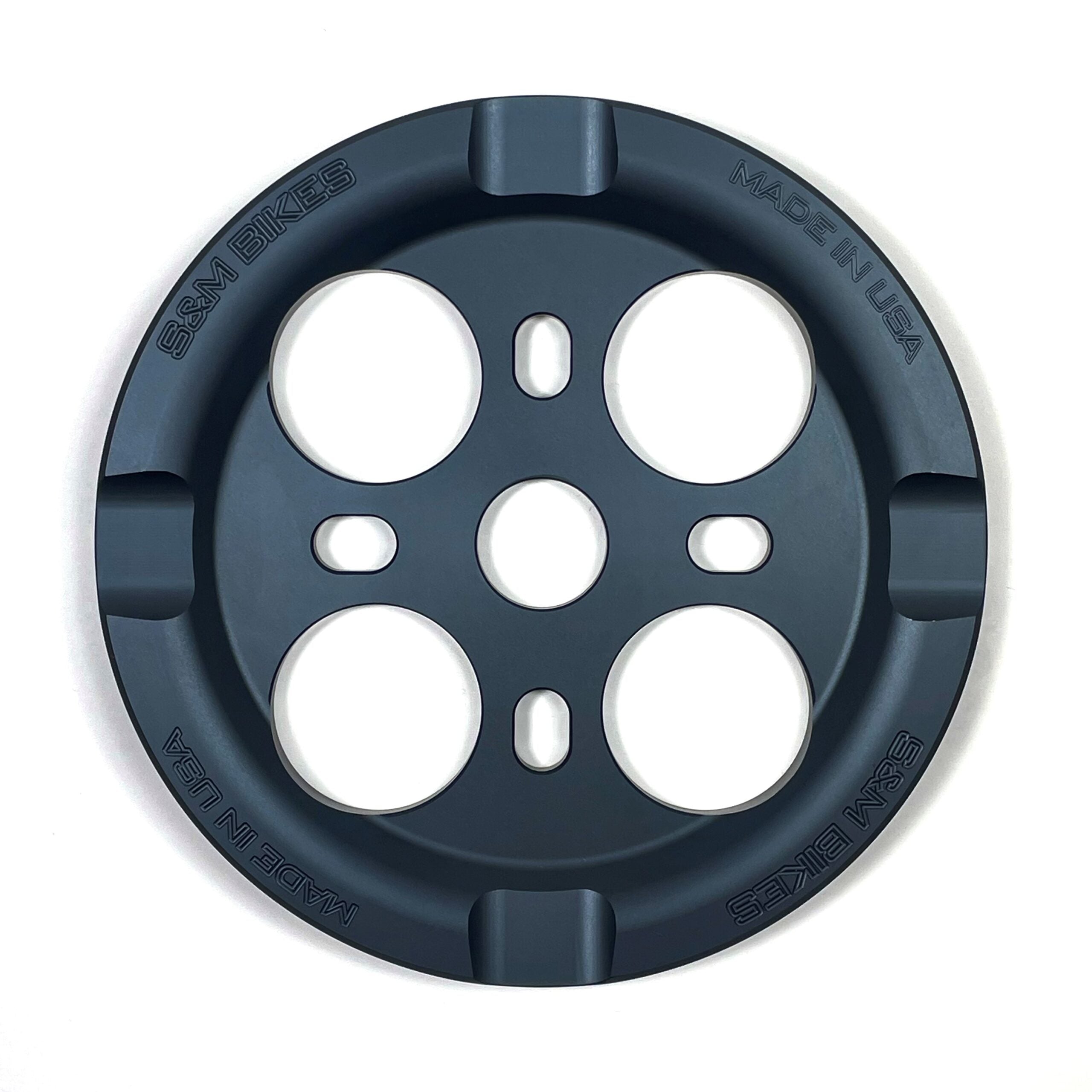 The S&M Mid School Guard Sprocket is a round black metal bashguard sprocket with four large holes, "S&M BIKES" and "MADE IN USA" engraved, designed by Aryei Levenson for added protection and style.