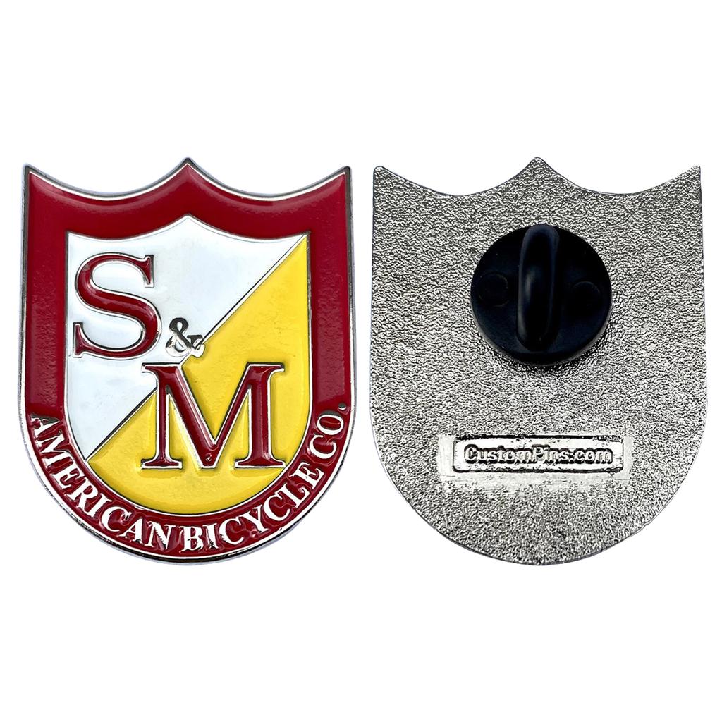 Front and back views of the S&M Shield Pin show an enamel shield design with "S & M" and "American Bicycle Co." on the front, while the back has a black rubber clutch and "CustomPins.com" marking.