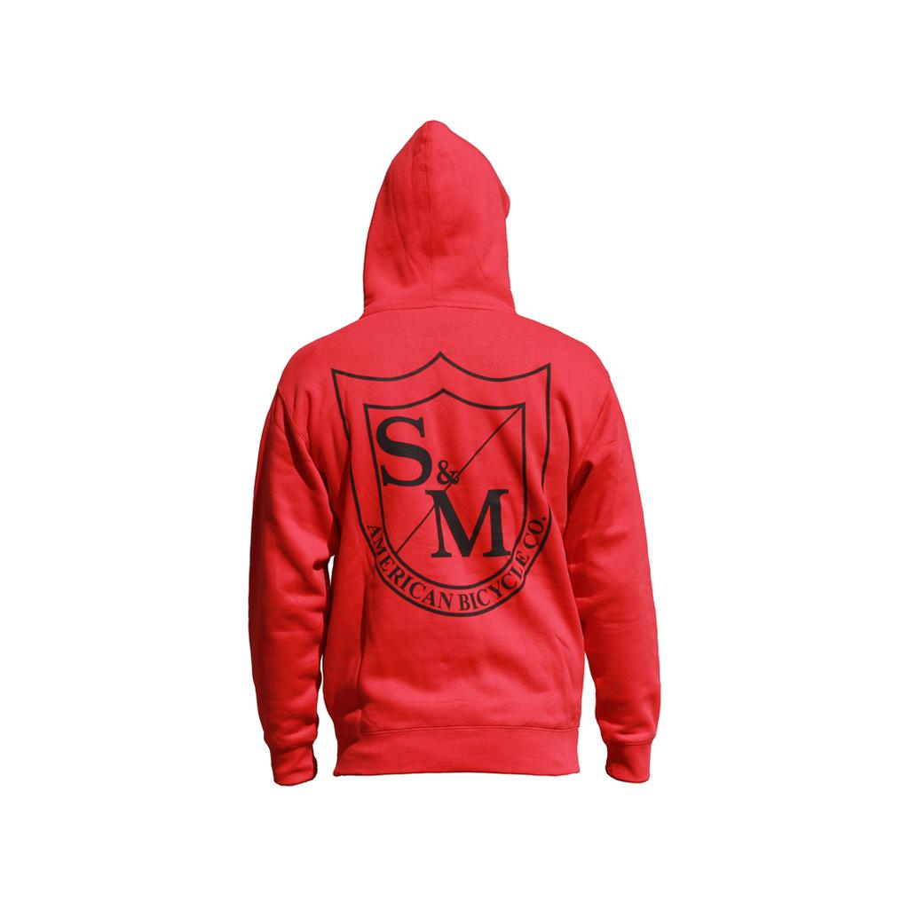 A person faces away from the camera wearing the S&M Two Shield Pullover Hoodie in red with a large black "S&M American Bicycle Co." logo and shield graphic on the back, featuring a cozy fleece-lined hood.