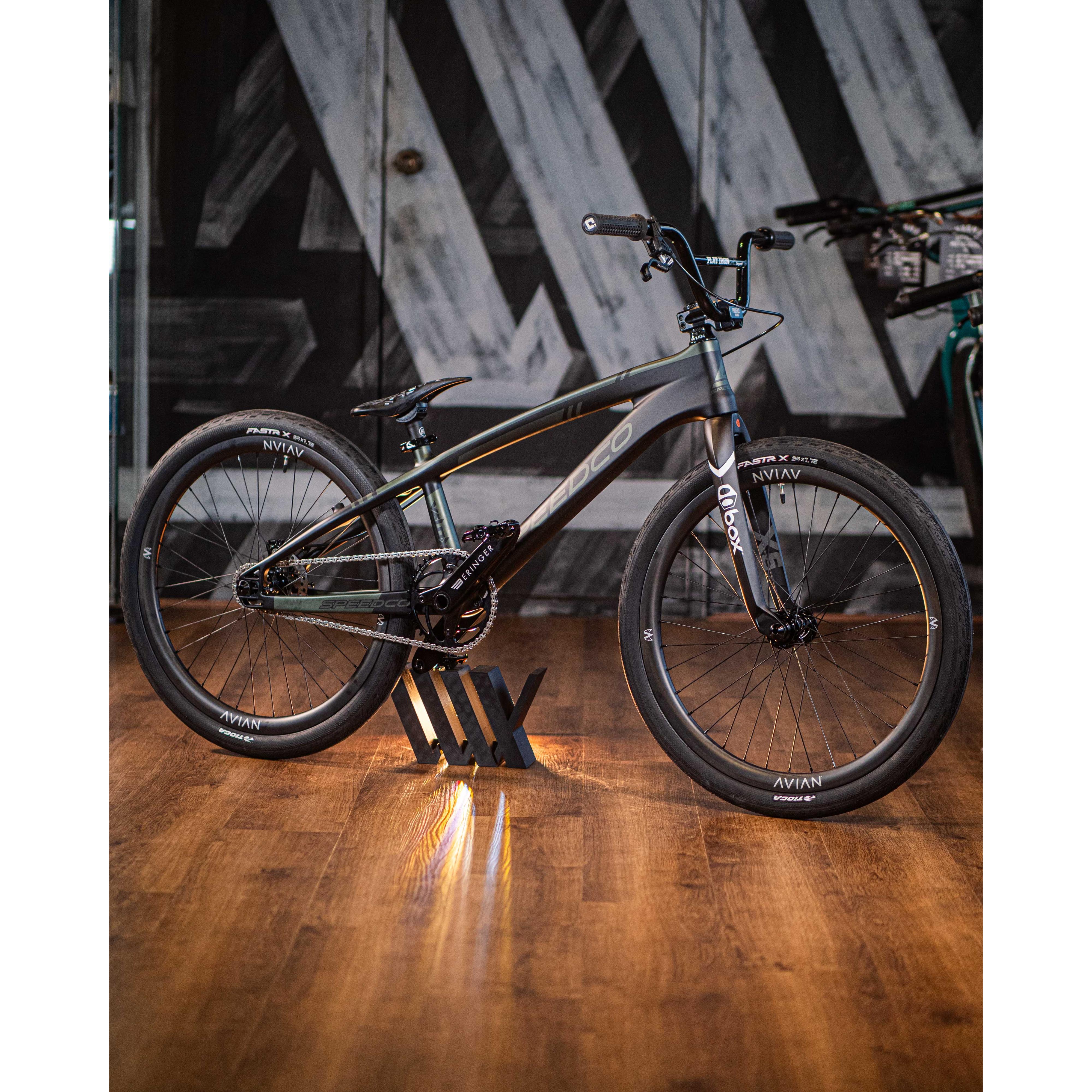A sleek black Speedco Velox Pro XL Cruiser Custom Bike, featuring its distinctive angled top tube, is showcased indoors on a wooden floor against a backdrop of geometric wall patterns.