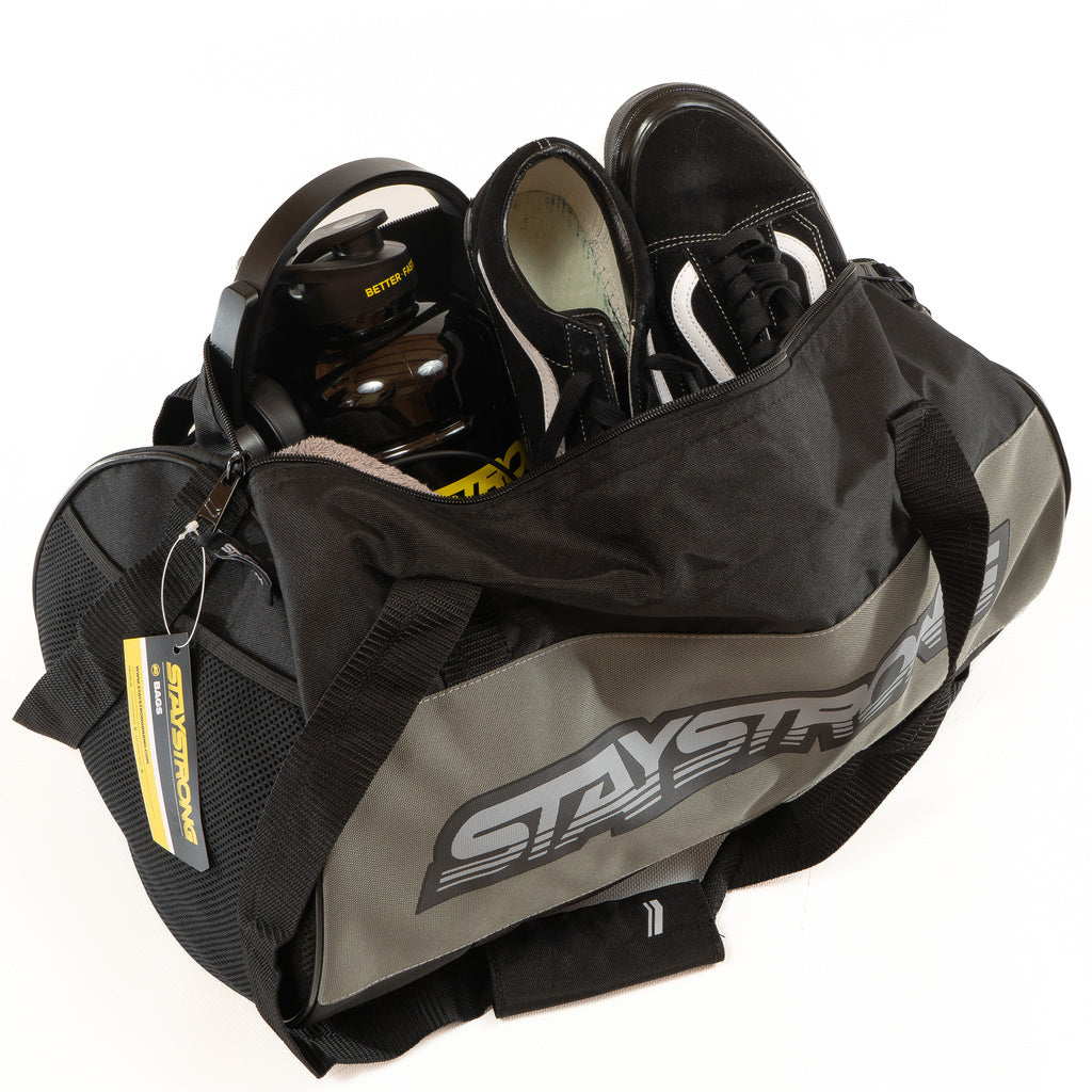 A Staystrong Duffle Bag Word Charcoal features “STAY STRONG” labeling and holds essential workout gear, including black athletic shoes, a water bottle, and a yellow-labeled accessory.