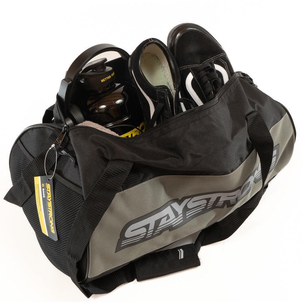 A Staystrong Duffle Bag Word Charcoal features “STAY STRONG” labeling and holds essential workout gear, including black athletic shoes, a water bottle, and a yellow-labeled accessory.