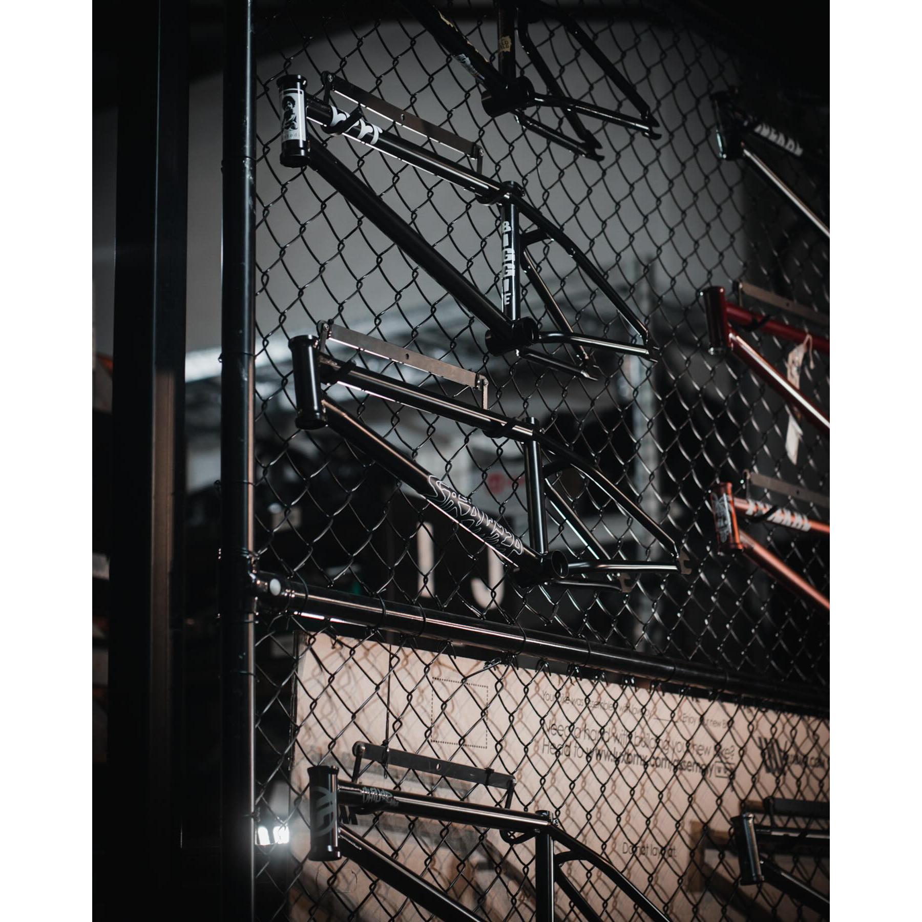 Bicycle frames in various colors, including the Stranger Royale Frame with integrated chain tensioners, are displayed on a black chain-link fence.