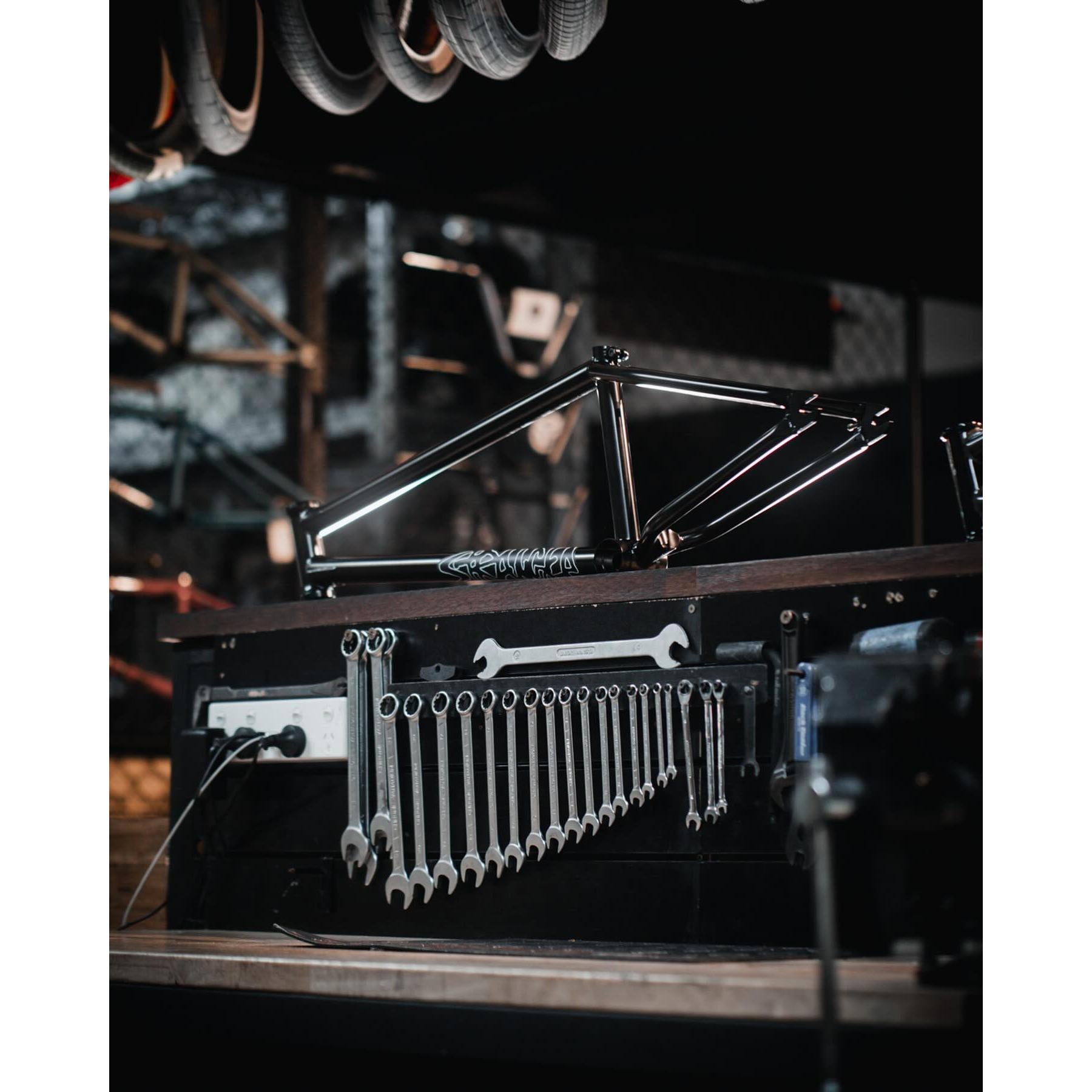 A bike frame, the Stranger Royale Frame, rests on a workbench with various wrenches neatly organized below it. Made from durable Chromoly tubing and featuring integrated chain tensioners, it sits ready for assembly. Overhead, several tires hang from the ceiling.