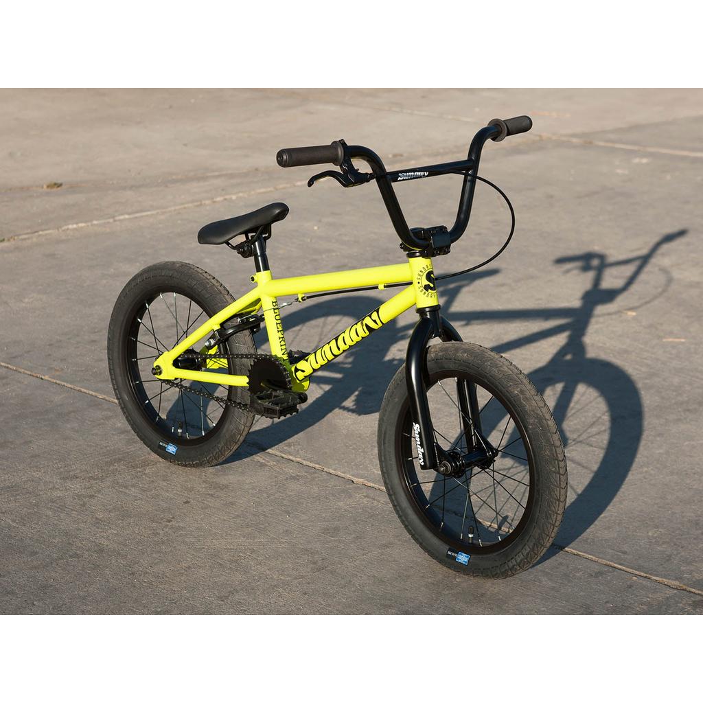 A Sunday Blueprint 16 Inch Bike, featuring a yellow frame, black handlebars and wheels, plus Odyssey Springfield Brakes, is parked on a concrete surface with its shadow visible.