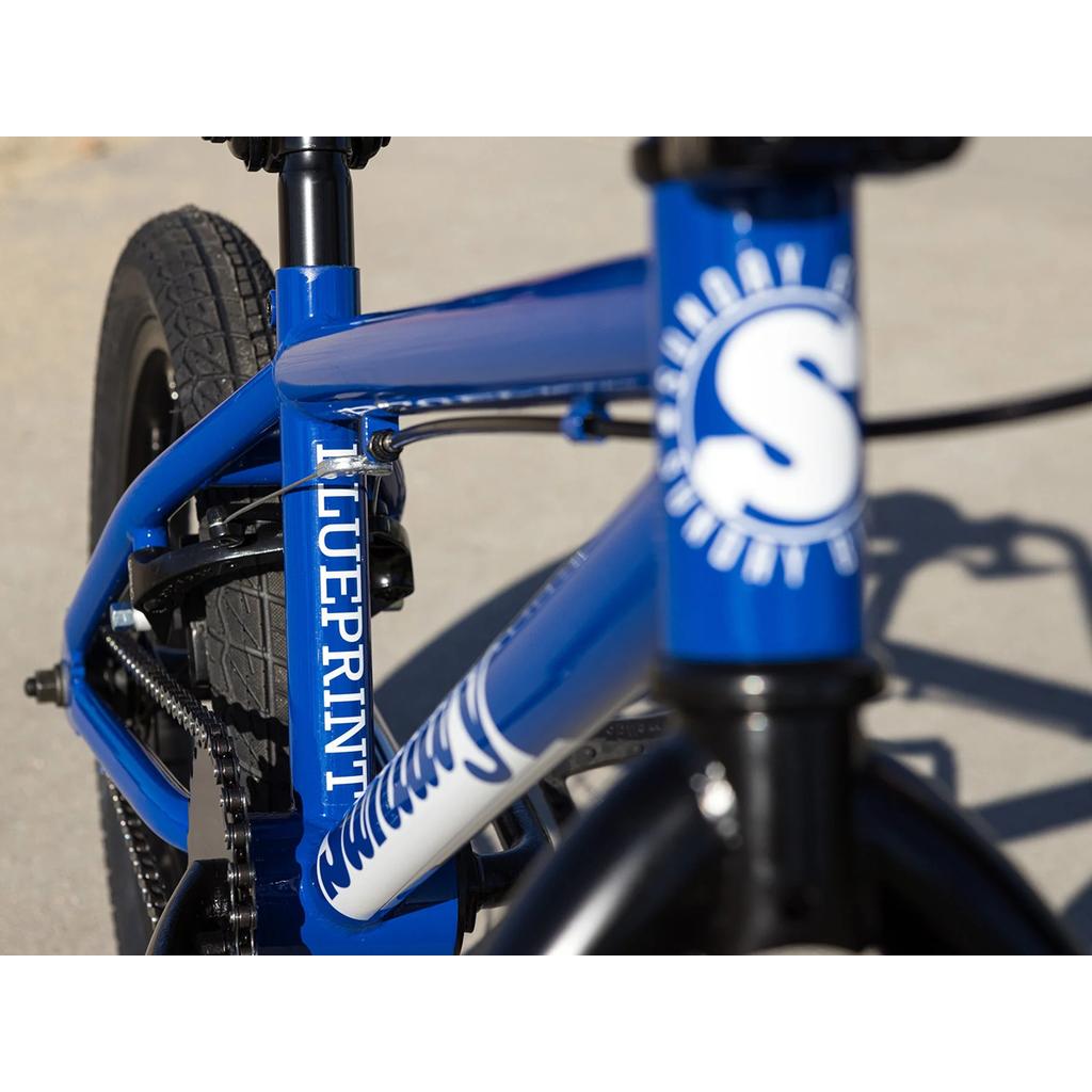 Close-up of the Sunday Blueprint 16 Inch Bike shows its blue frame with "BLUEPRINT" on the tube and a bold white "S" logo on the front, highlighting classic BMX geometry. The bike is pictured outdoors on a paved surface.