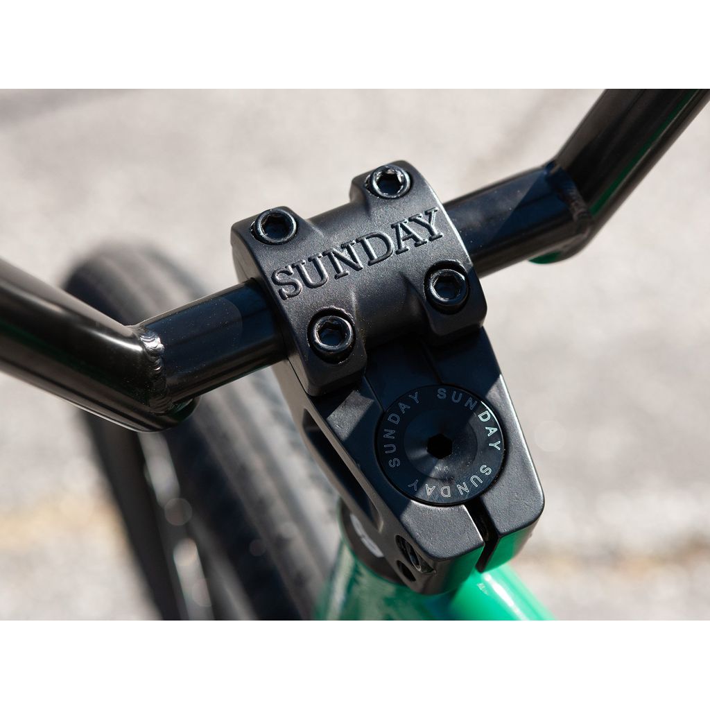 Close-up of a black bike stem with the brand name "Sunday" etched on it, connecting the handlebars. The Sunday Forecaster 20 Inch Bike (2022) / Alec Siemon's green frame and tires are also partially visible, boasting a 100% chromoly frame for durability.