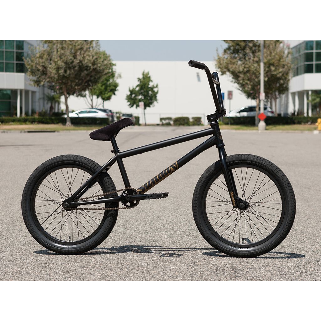 A sleek black Sunday Forecaster Broc Raiford 20 Inch Bike (2023) with a chromoly frame and thick tires is parked on an outdoor asphalt surface, featuring modern buildings and greenery in the background.