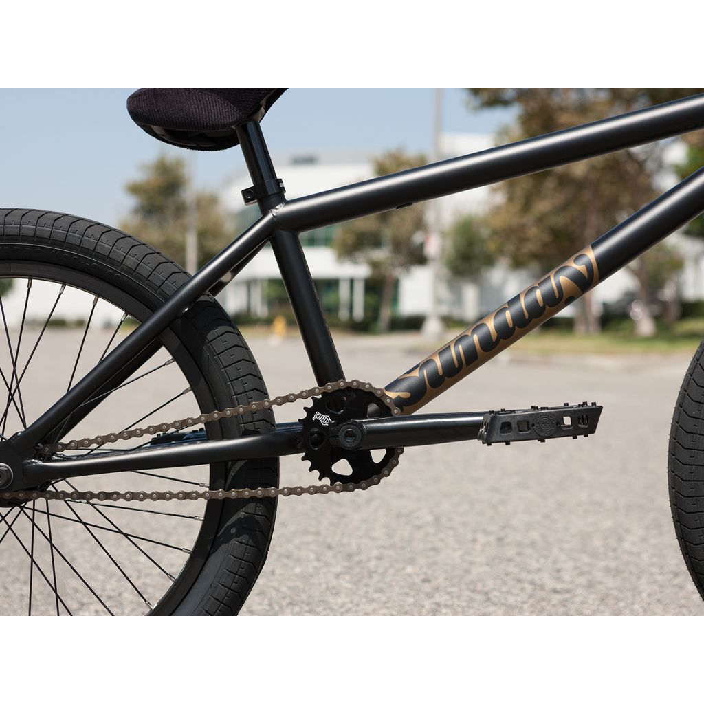 Close-up of the drivetrain and rear wheel of a black Sunday Forecaster Broc Raiford 20 Inch Bike (2023), highlighting the chain, pedals, and sprockets on its robust chromoly frame. The background shows blurred trees and a building.