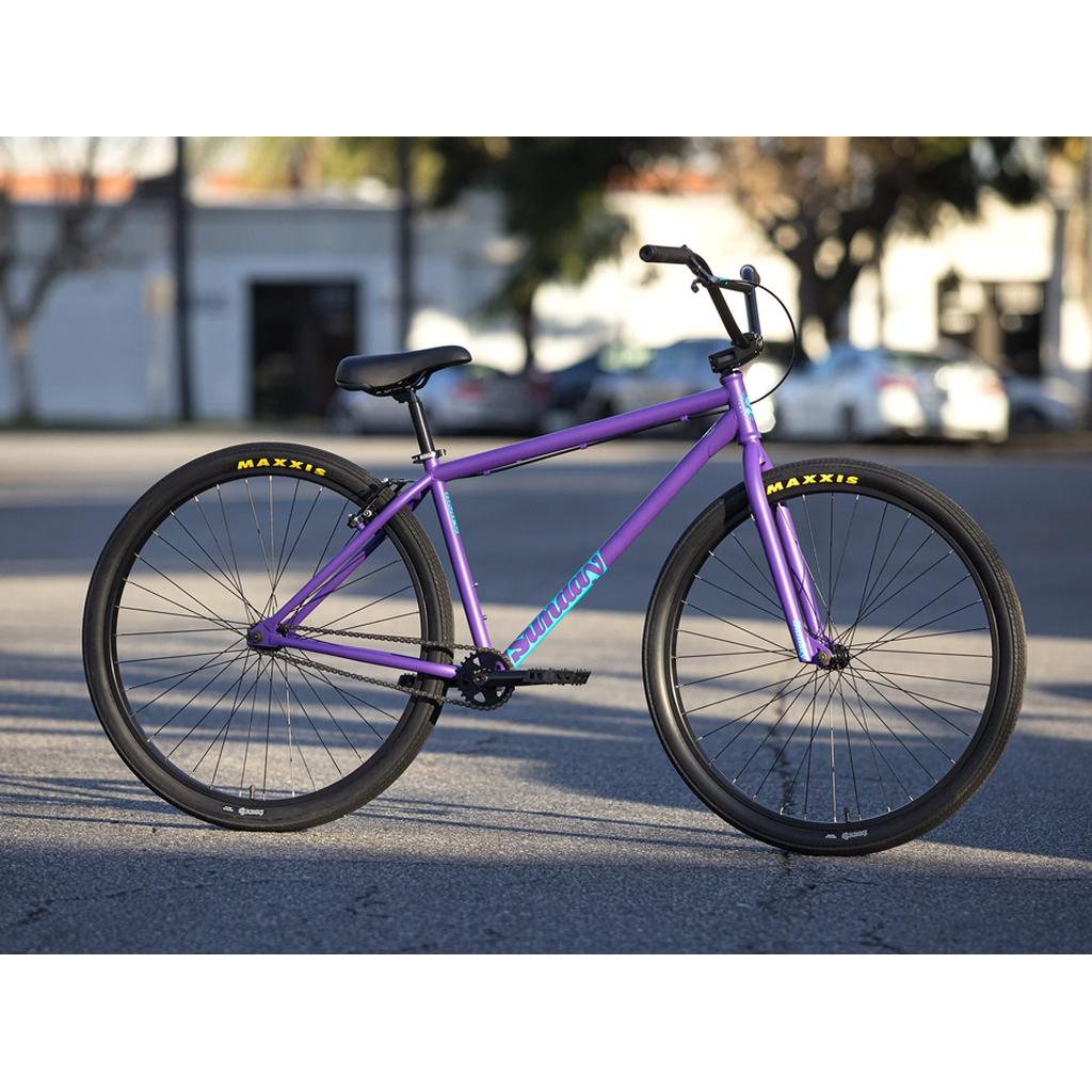 A Sunday High C 29 Inch Bike (2025) with a purple chromoly frame, single-speed drivetrain, black handlebars, and Maxxis tires is parked on a paved street with buildings and trees in the background.