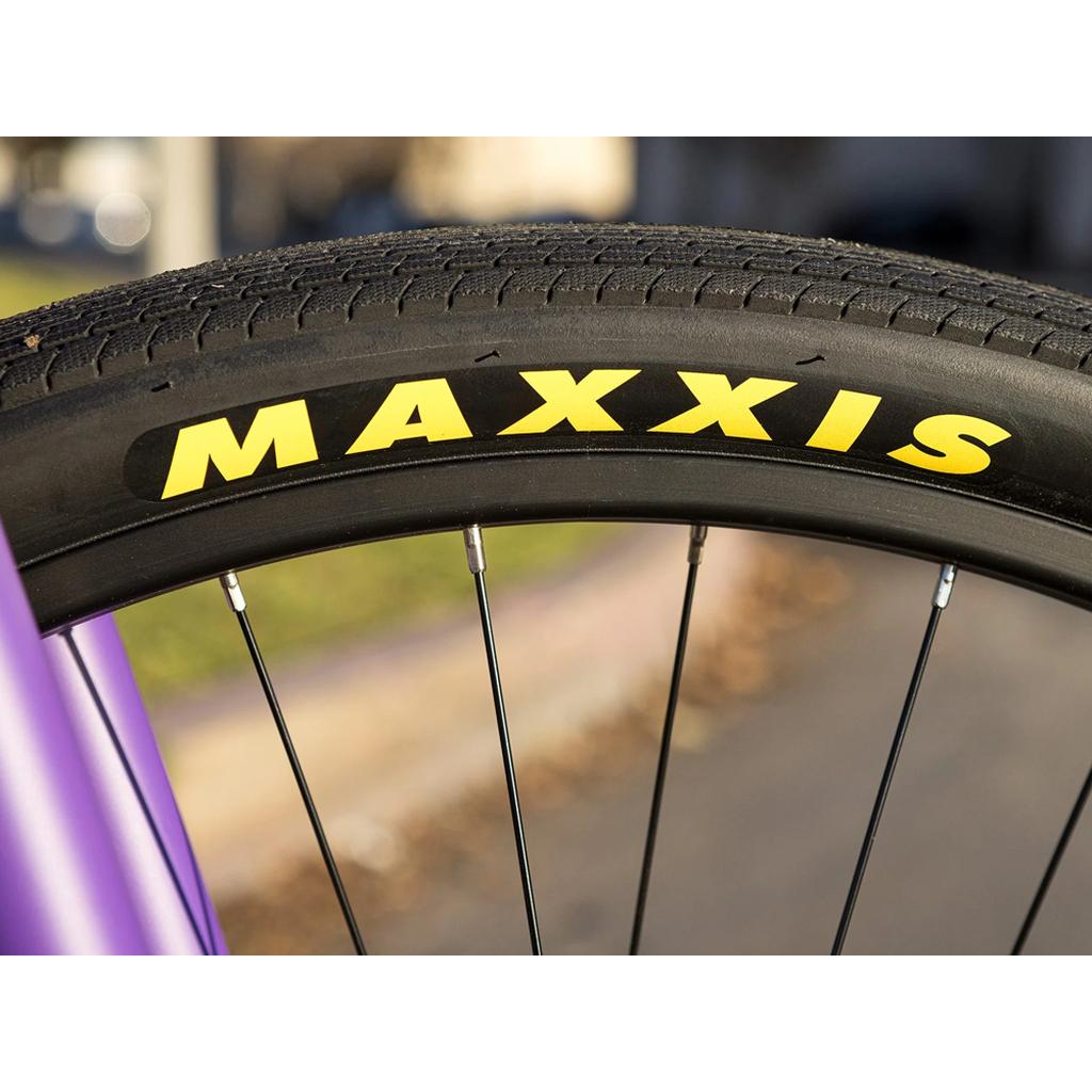 Close-up of a bicycle wheel with a black tire displaying yellow "MAXXIS" text, set against a blurred backdrop highlighting the modern geometry of the 2025 Sunday High C 29 Inch Bike.