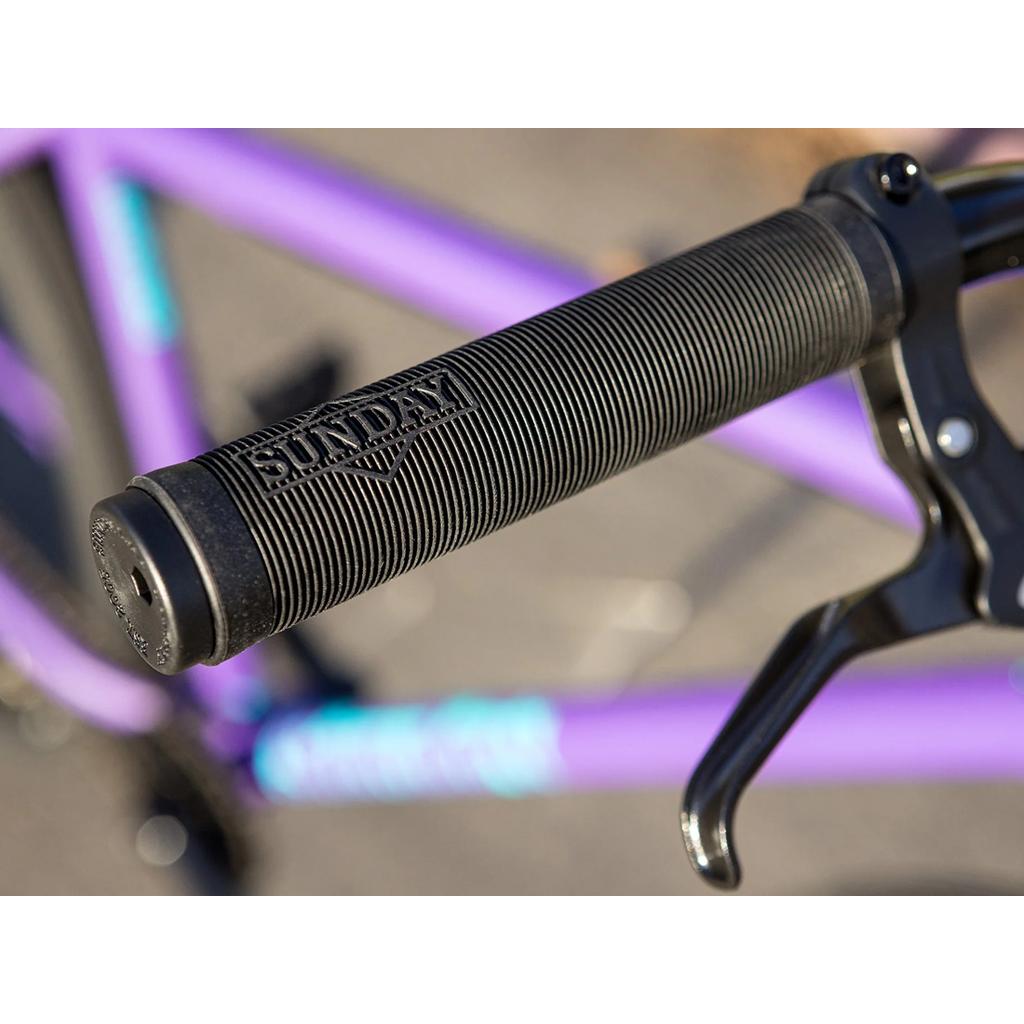 Close-up of a black handlebar grip with "SUNDAY" branding on the Sunday High C 29 Inch Bike (2025) in purple, showcasing its geometry and a right brake lever.