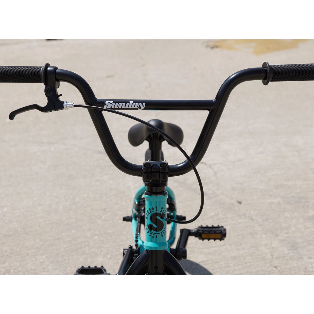 A close-up view of the handlebars and front of a blue Sunday Primer 16 Inch Bike (2025) on a concrete surface. "Sunday" is printed on the handlebars, showcasing its Odyssey Springfield Brakes.