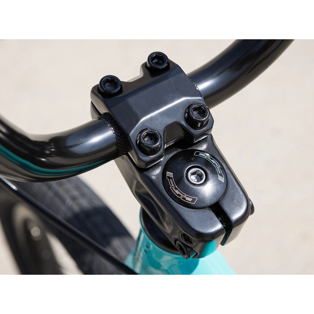 Close-up of a black bicycle handlebar and stem with four bolts and a cap on the Sunday Primer 16 Inch Bike (2025). The teal bike frame stands out against the gray pavement in the background, showcasing precision components like the Odyssey Springfield Brakes.
