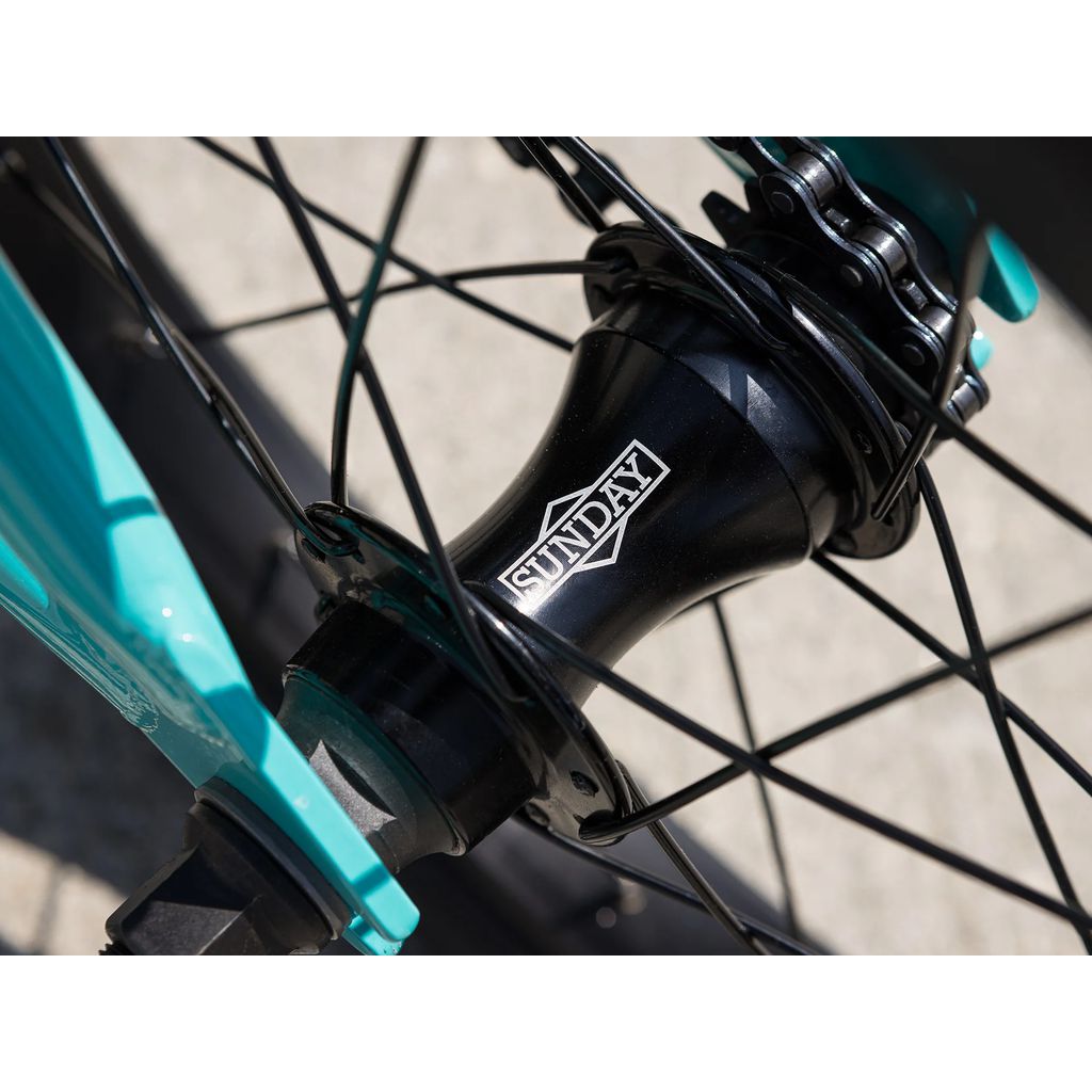 Close-up of a BMX bike wheel hub bearing the "Sunday" logo. The black hub and spokes connect to a teal frame. The wheel, which is part of the 2025 Sunday Primer 16 Inch Bike, rests on a gray concrete surface.