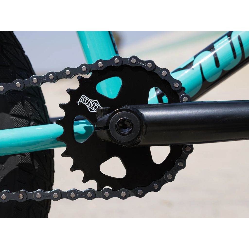 Close-up image of a black chainring and crankarm on the Sunday Primer 16 Inch Bike (2025), attached to a teal frame with Odyssey Springfield Brakes and a section of the tire visible in the background.