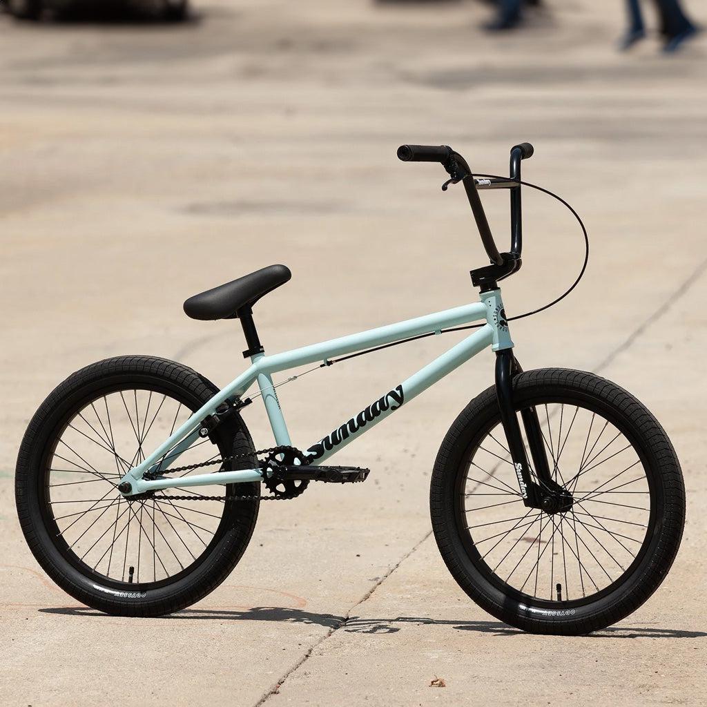 A Sunday Primer 20 Inch Bike, painted in light blue with black handlebars and tires, showcasing the "Sunday Primer" logo on the frame, is parked on a paved surface. This starter bike offers pro-level geometry for an optimal riding experience.