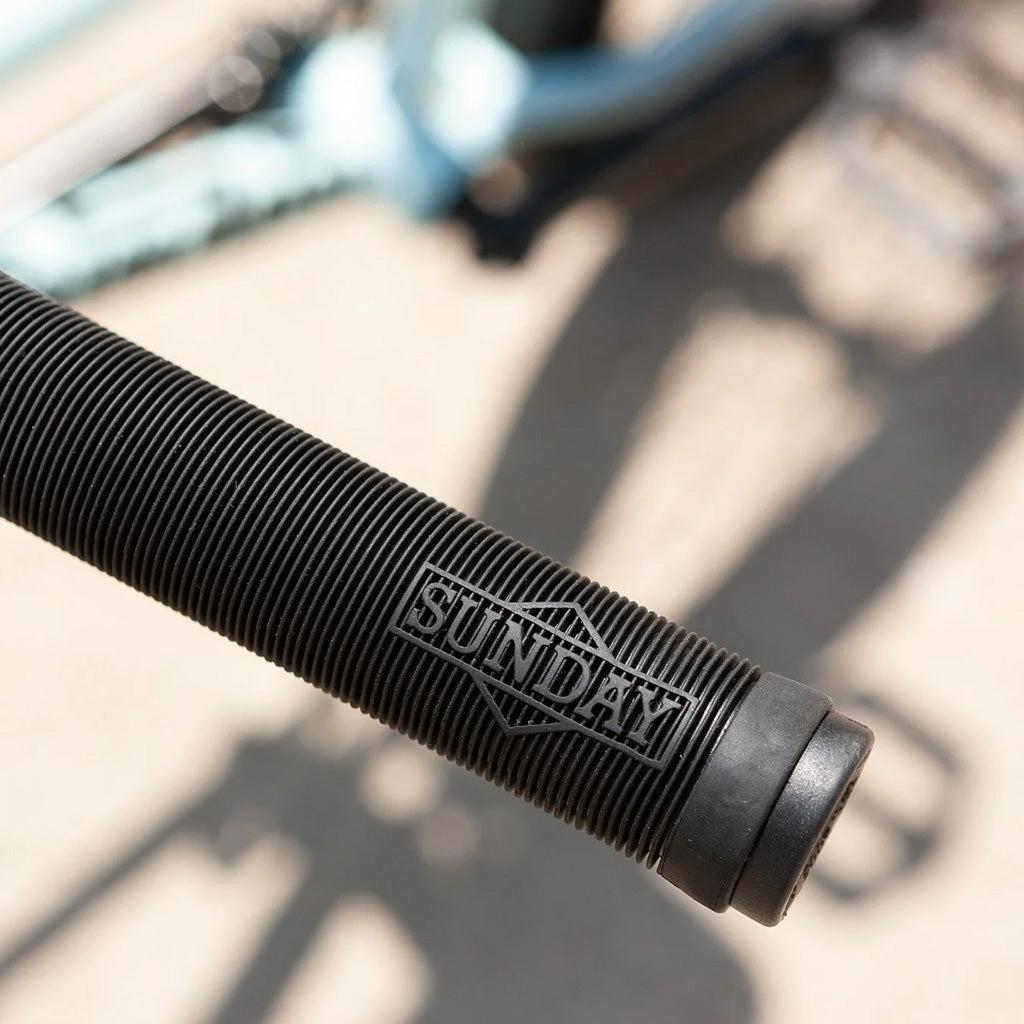 Close-up of a black bike handlebar grip with the brand "Sunday" embossed on it, showcased on a starter bike featuring pro-level geometry. The bicycle in the blurred background hints at the Sunday Primer 20 Inch Bike model.