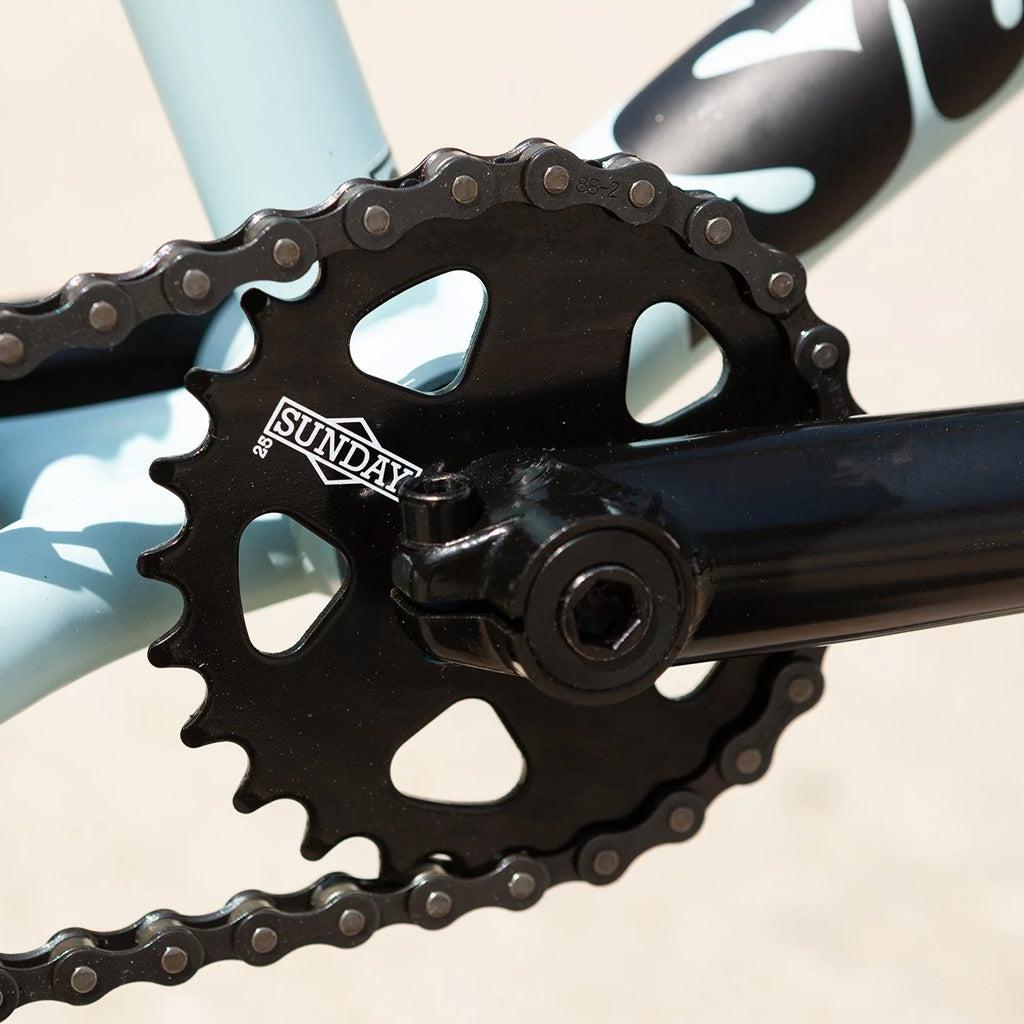 Close-up image of a black bicycle crankset and chainring with the brand name "Sunday" visible on the chainring. The light blue frame of the Sunday Primer 20 Inch Bike is partially visible in the background.