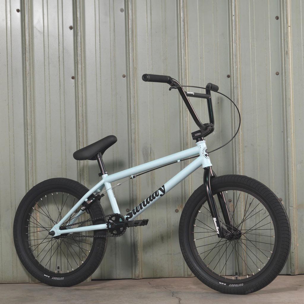 A Sunday Primer 20 Inch Bike in light blue, featuring black handlebars, seat, and tires, is positioned against a corrugated metal wall. The starter bike, boasting pro-level geometry, proudly displays "Sunday" on its frame.