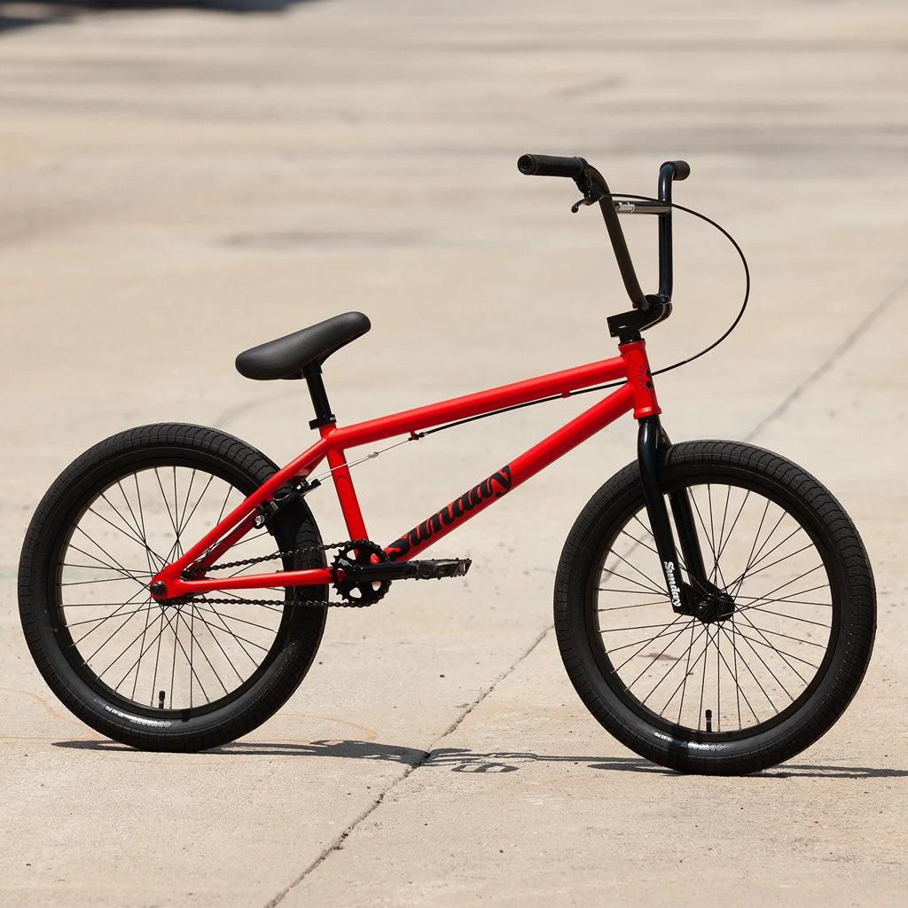 A Sunday Primer 20 Inch Bike, featuring a red frame with black handlebars and tires, is parked on a concrete surface. Its frame showcases a simple design and pro-level geometry, equipped with a front brake cable.