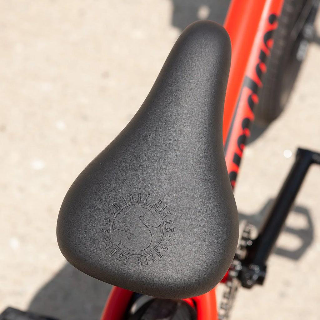Close-up of a black bicycle seat on a red Sunday Primer 20 Inch Bike frame, with "Sunday Bikes" embossed on the seat.