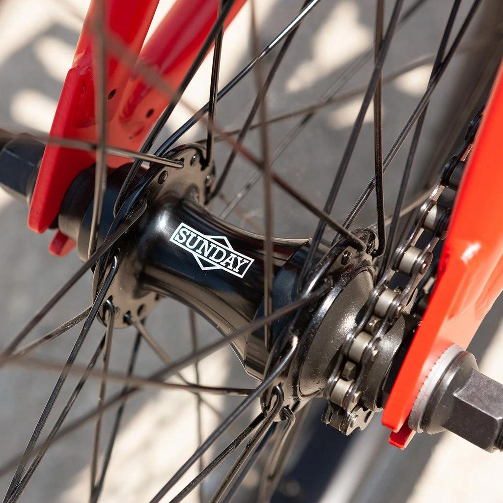 Close-up of the bicycle wheel hub with spokes radiating outward. The hub, part of the Sunday Primer 20 Inch Bike, has the brand name "Sunday" printed on it. The bicycle frame is red, showcasing its pro-level geometry.