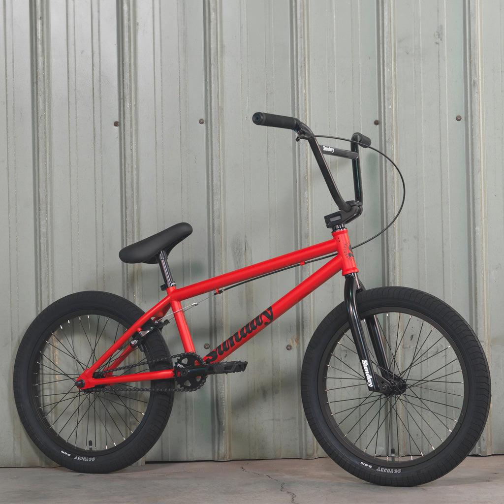A Sunday Primer 20 Inch Bike, featuring a red frame with black handlebars and tires and boasting pro-level geometry, is parked against a corrugated metal wall.