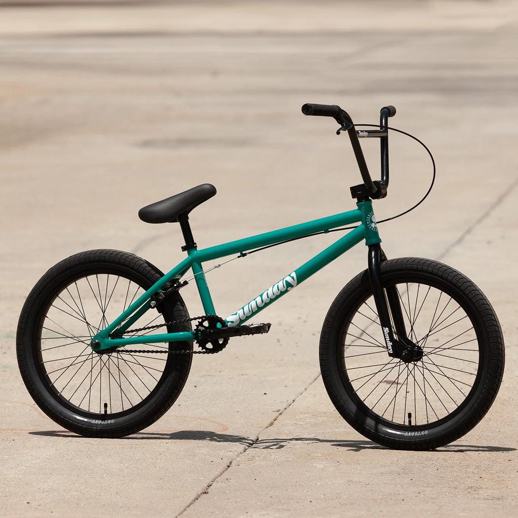 A teal Sunday Primer 20 Inch Bike, designed with pro-level geometry and featuring black handlebars and black tires, is parked on a concrete surface.