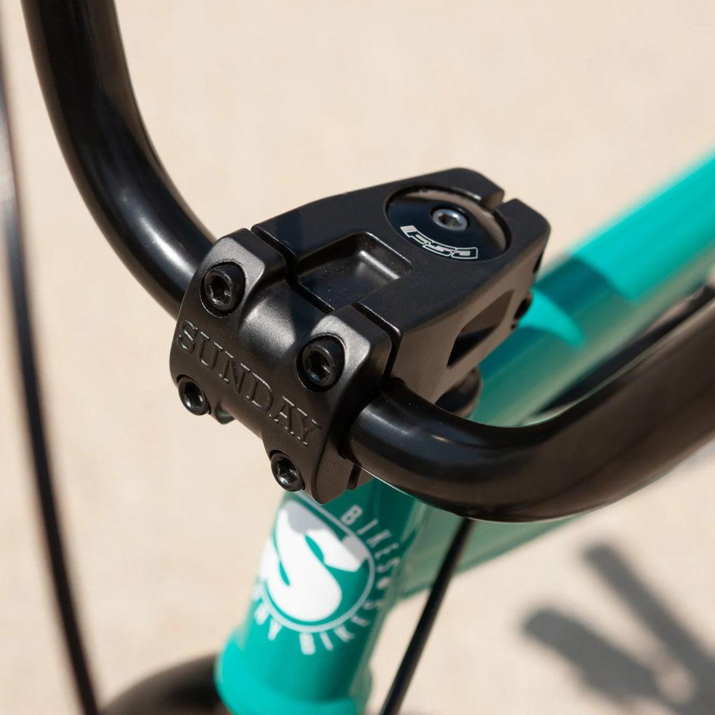 Close-up of a black handlebar and stem on a teal Sunday Primer 20 Inch Bike. The word "SUNDAY" is engraved on the front of the stem, showcasing its pro-level geometry.