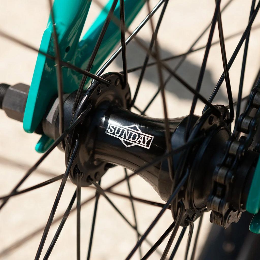 A close-up of the Sunday Primer 20 Inch Bike wheel hub features black spokes and part of the green frame, showcasing pro-level geometry ideal for a starter bike.
