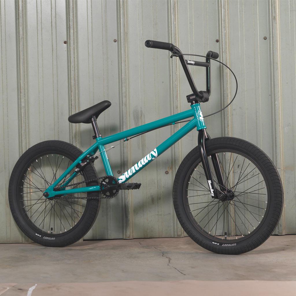 A teal Sunday Primer 20 Inch BMX bike, featuring black wheels and handlebars, is positioned against a corrugated metal wall. The brand name "Sunday Primer" is prominently displayed on the frame, highlighting its pro-level geometry ideal for a starter bike.