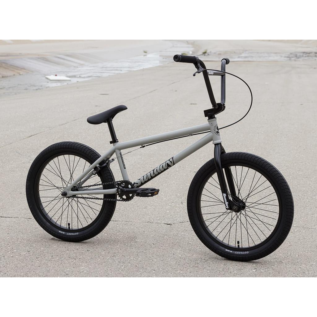 A gray Sunday Primer 20 Inch Bike with black handlebars, seat, and tires rests on concrete near a dry channel—an ideal BMX pick for beginners.