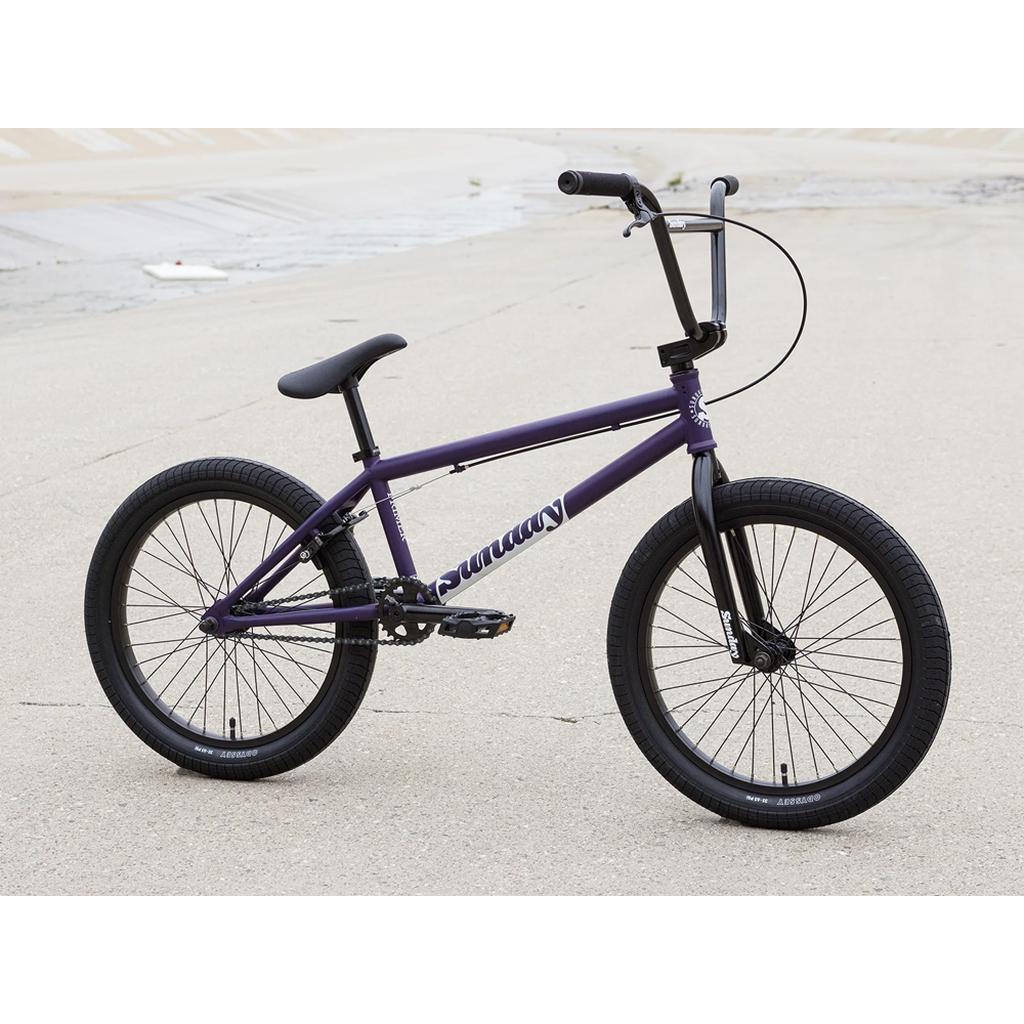 A purple Sunday Primer 20 Inch Bike with black wheels, pedals, and handlebars is parked on concrete by a drainage canal—a great beginner’s BMX bike featuring the reliable Odyssey Springfield U-brake.