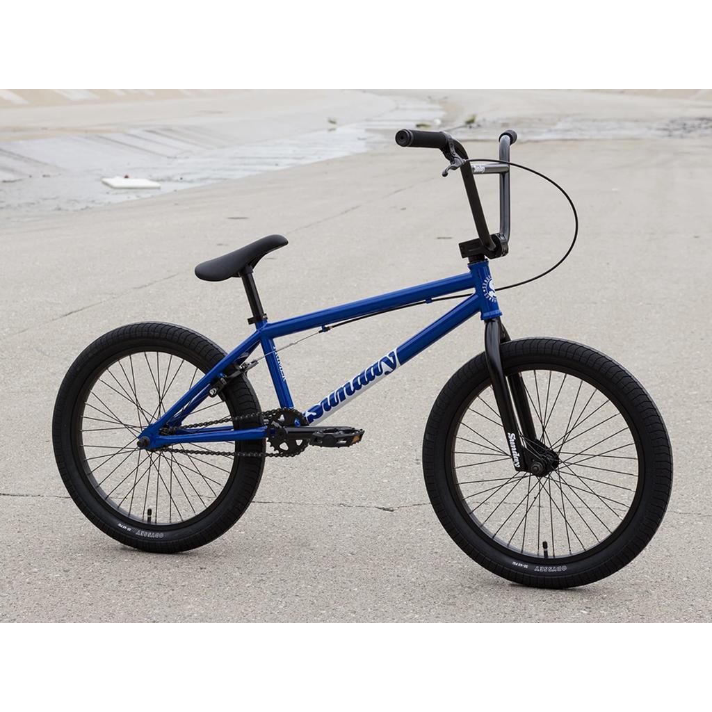 A Sunday Primer 20 Inch Bike in blue with black wheels and Odyssey Springfield handlebars is parked on a paved surface near a concrete waterway.