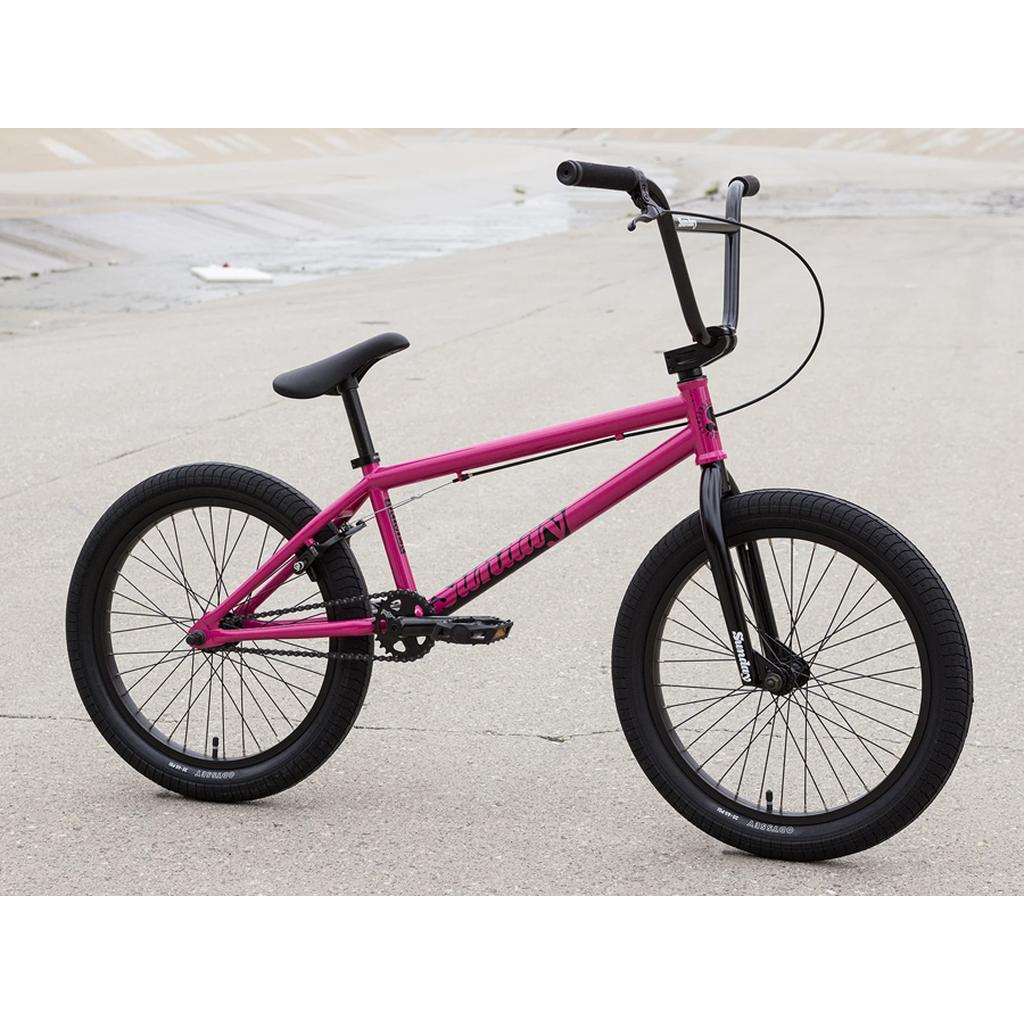 A magenta Sunday Primer 20 Inch Bike with black tires, seat, and Odyssey Springfield handlebars is parked on concrete near a drainage channel.