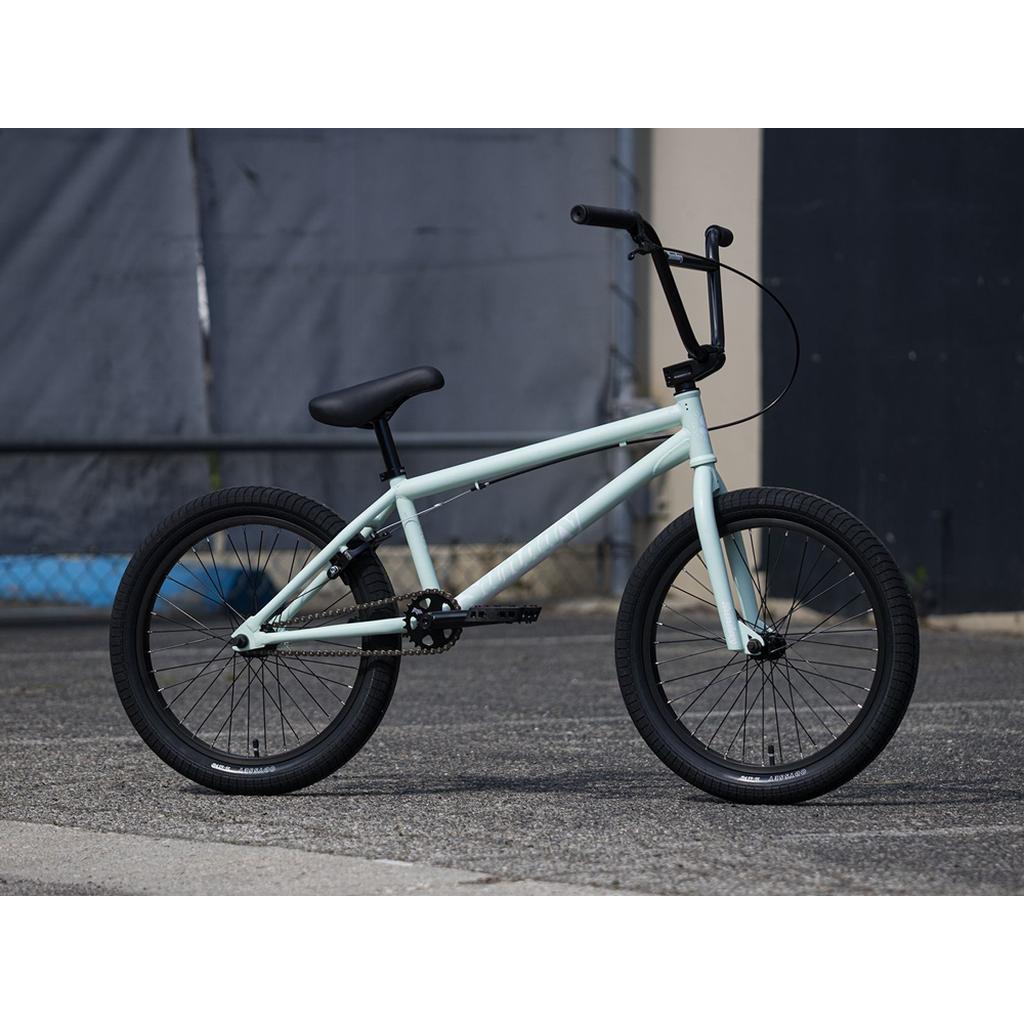 A Sunday Scout 20 Inch Bike in light blue with black Odyssey handlebars, seat, and tires is parked on a paved surface outdoors near a building.