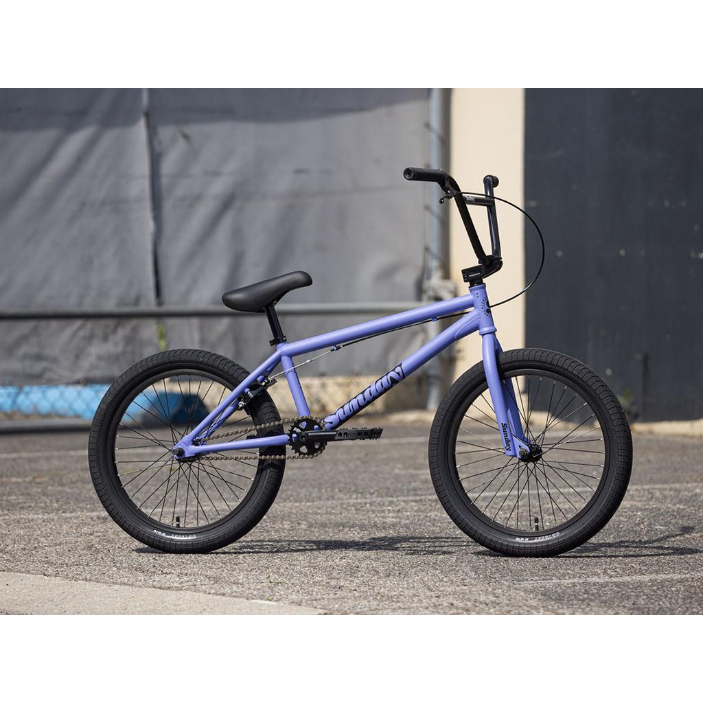 A Sunday Scout 20 Inch Bike in purple with black Odyssey handlebars and tires is parked outside on pavement near a building and a chain-link fence.