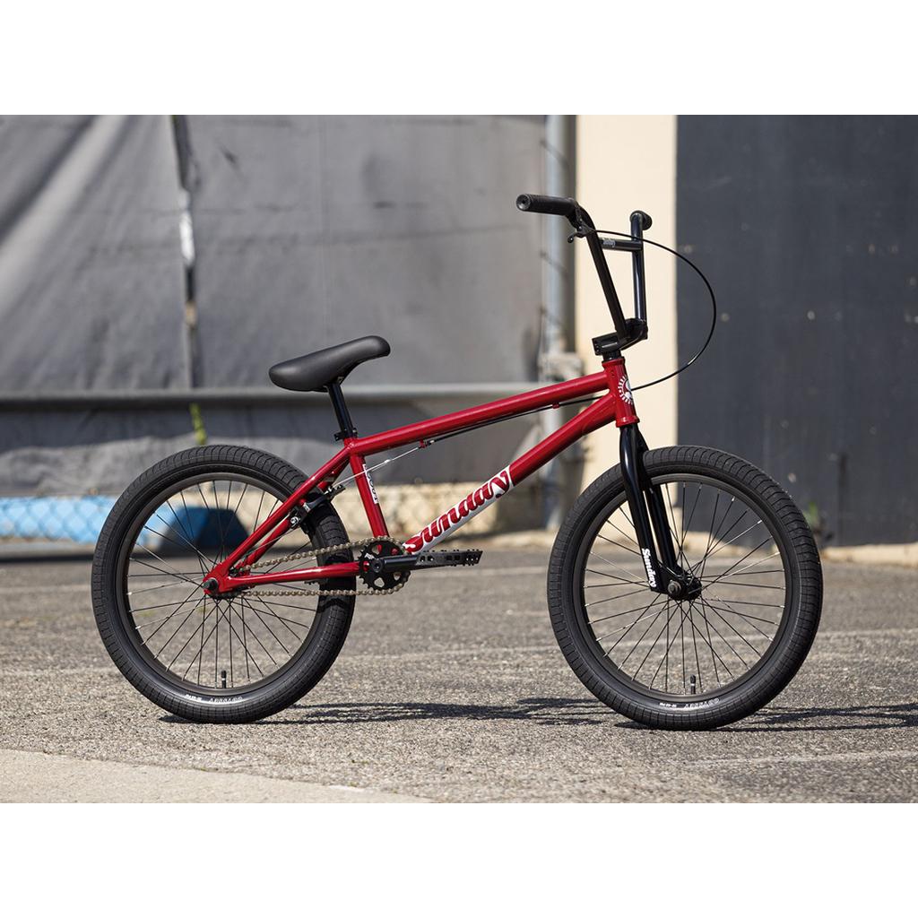 A red Sunday Scout 20 Inch Bike with black handlebars and tires is parked on pavement near a gray and beige building.