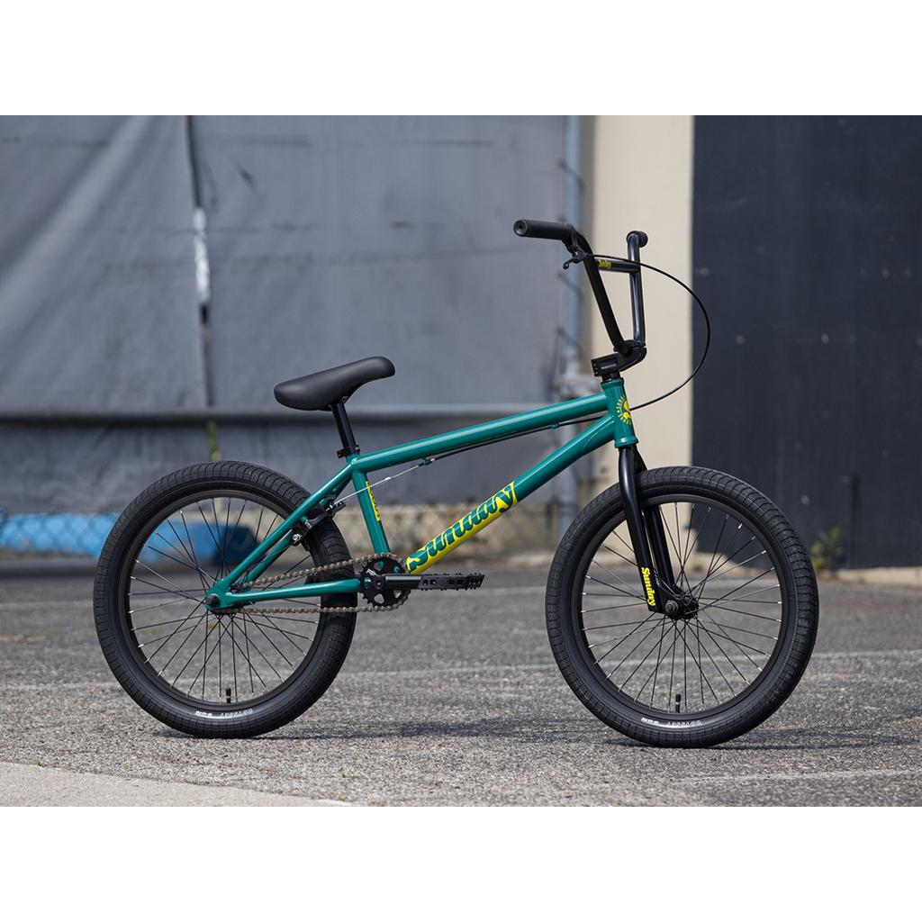 A teal Sunday Scout 20 Inch Bike with black Odyssey tires and handlebars is parked on a paved surface in front of a gray and cream-colored background.