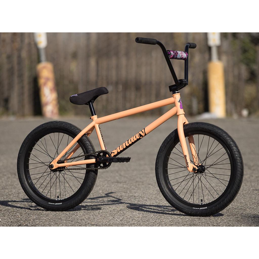 A peach Street Sweeper 20 Inch BMX bike with a Jake Seeley signature chromoly frame, black handlebars, and tires is parked on an asphalt surface near two yellow bollards.